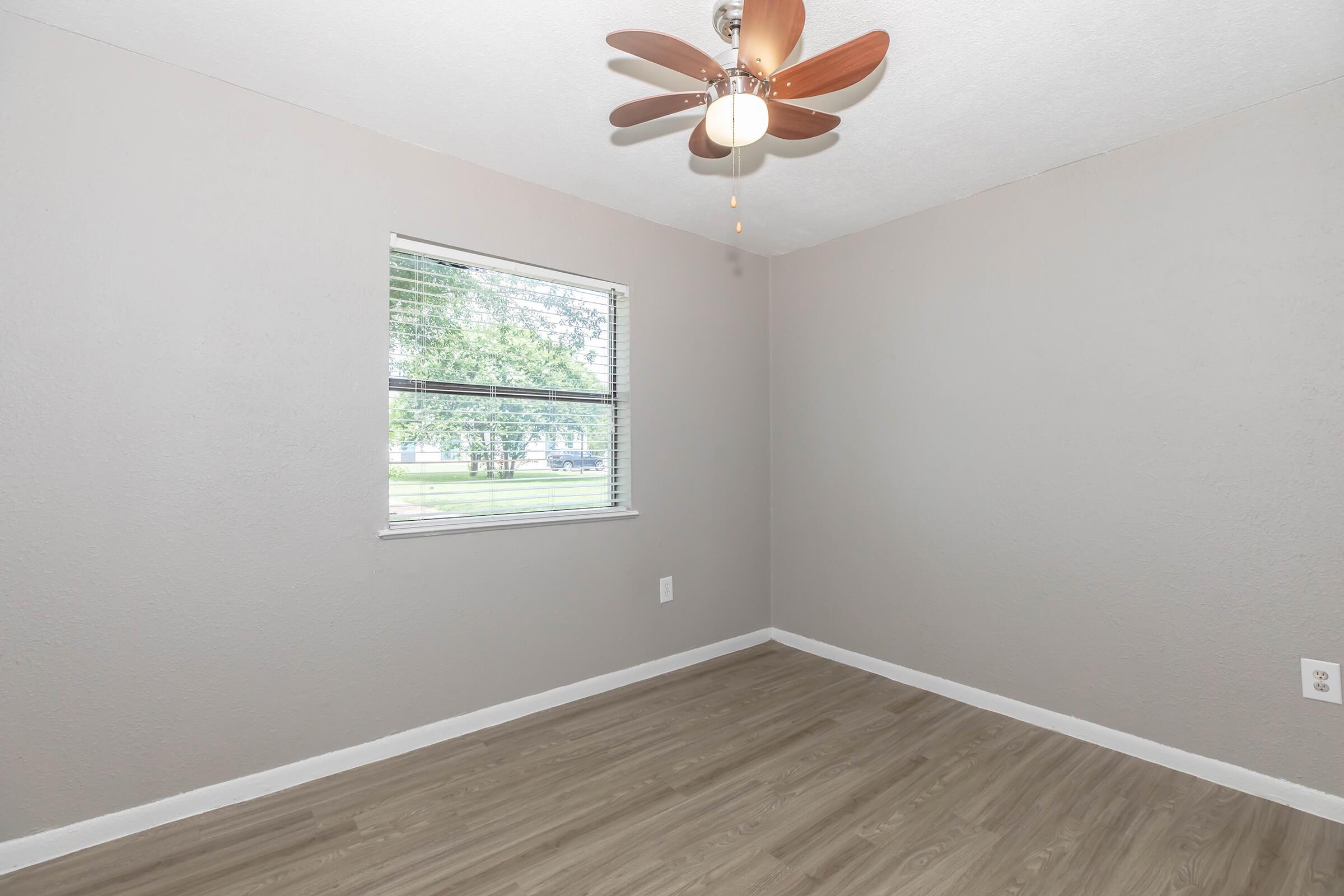 Empty room with light gray walls, a ceiling fan with wooden blades, and a laminate floor. A window with blinds lets in natural light, revealing greenery outside. The room has no furniture, creating a spacious, minimalistic appearance.