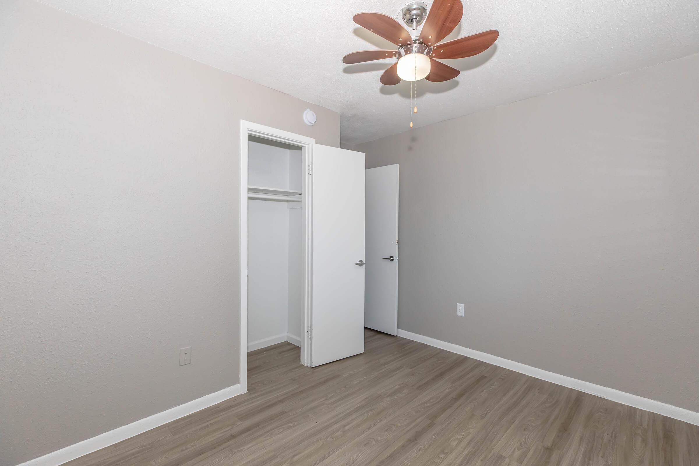 A clean, empty bedroom featuring light gray walls, a light wooden floor, and a ceiling fan with wooden blades. One door is open to a closet, while another door leads to an adjacent space, enhancing the room's spacious feel. Natural light illuminates the room, creating a bright atmosphere.