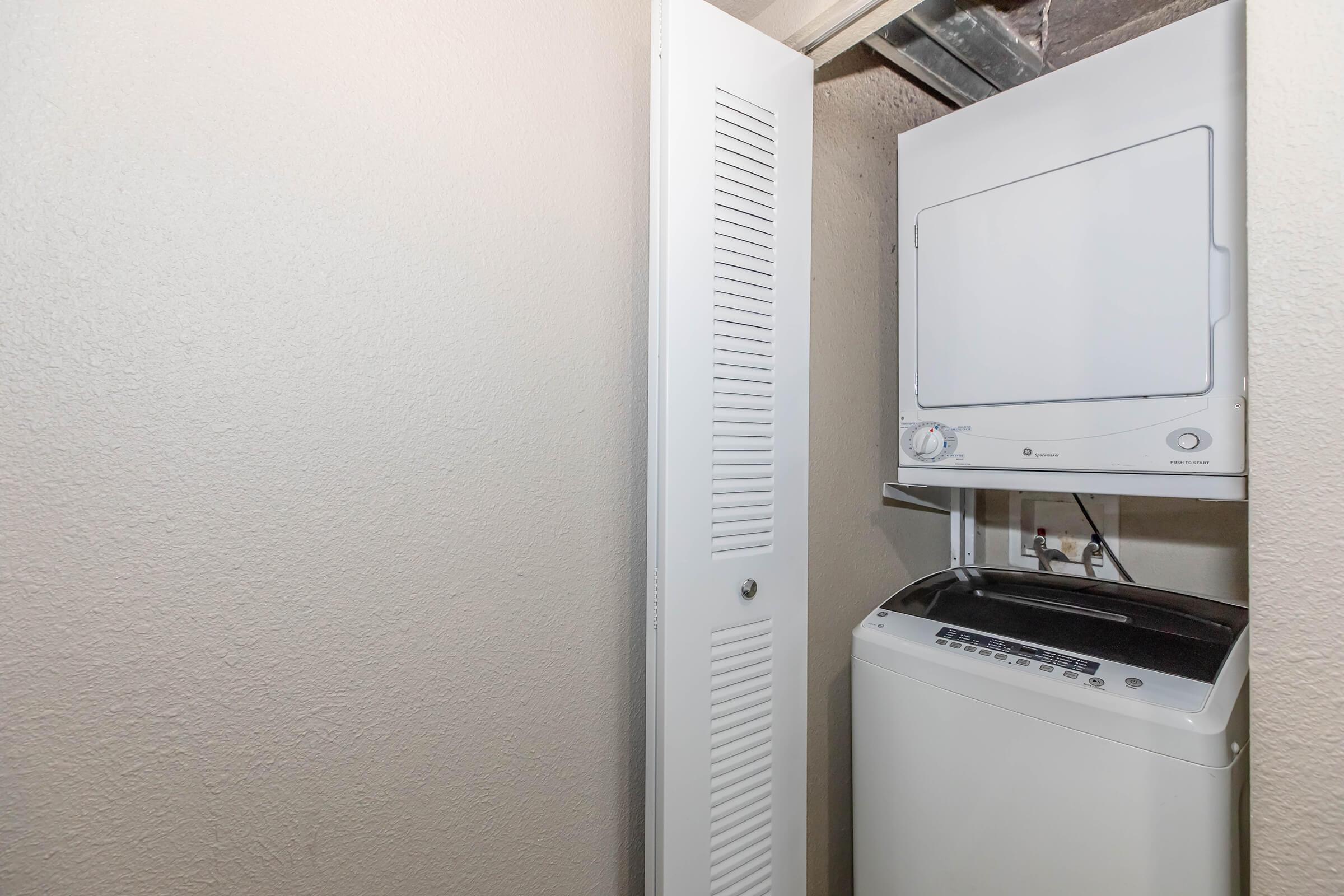 A compact laundry area featuring a stacked washer and dryer unit. The washer is on the bottom and the dryer on top, situated in a corner against a light-colored wall. A nearby door with vertical slats leads to additional storage space. The area is clean and organized.