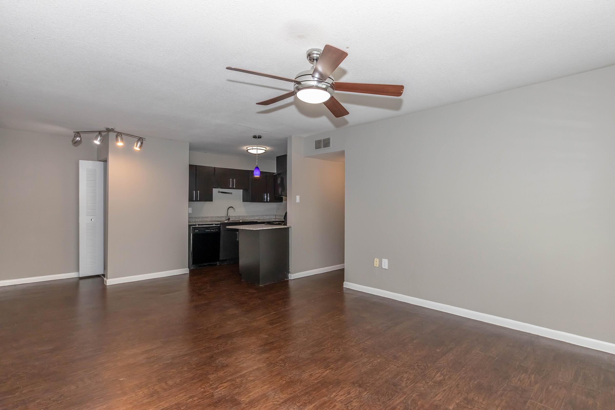 Spacious living area with brown laminate flooring, a ceiling fan with wooden blades, and a modern kitchen visible in the background. The kitchen features dark cabinetry and a sleek countertop. The walls are painted in a light gray color, offering a contemporary feel, with ample natural light.