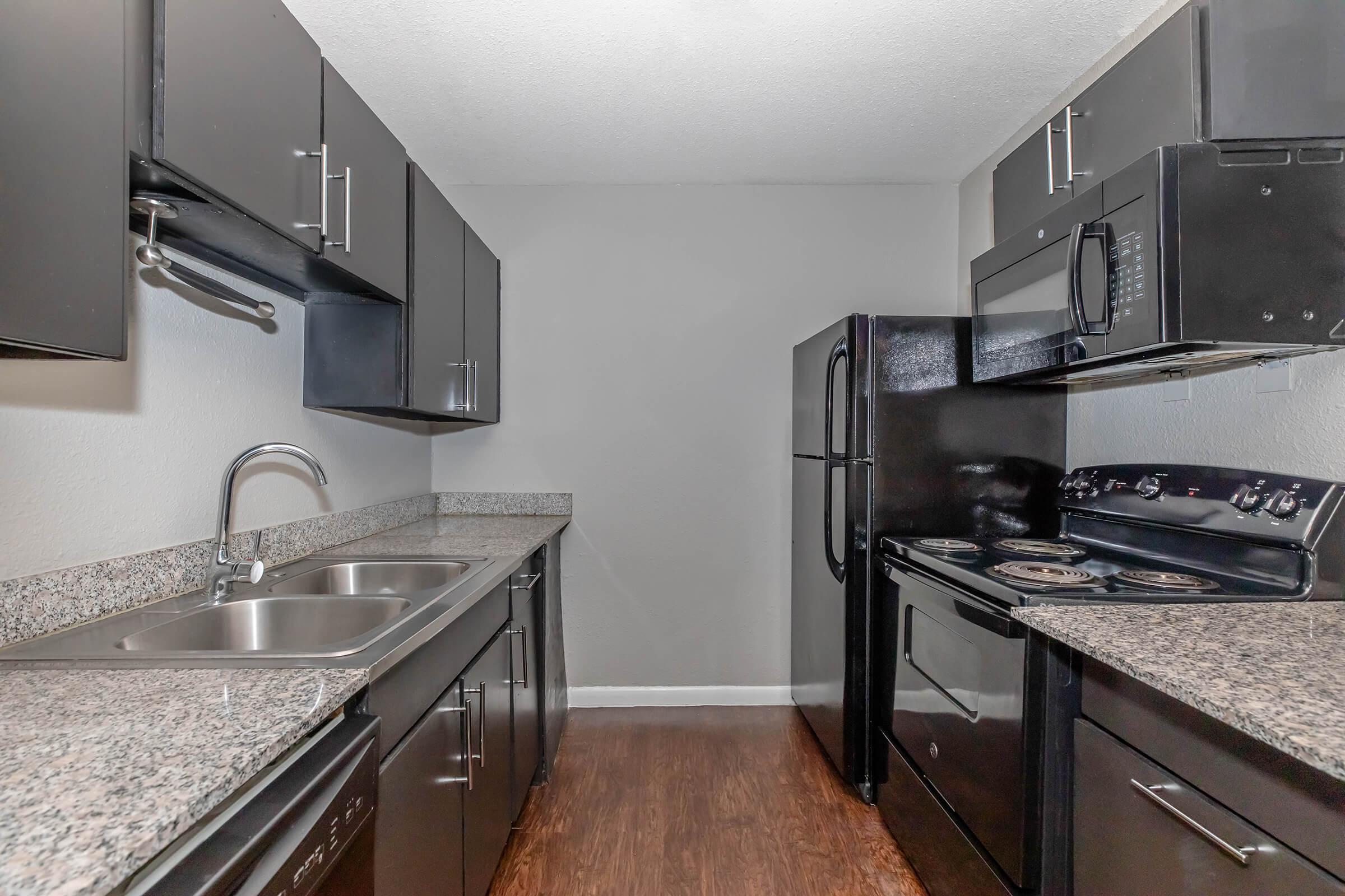 A modern kitchen with dark cabinets, granite countertops, a double sink, and stainless steel appliances including a stove, microwave, and refrigerator. The walls are painted gray, and the flooring is a dark wood laminate, creating a sleek and contemporary look.