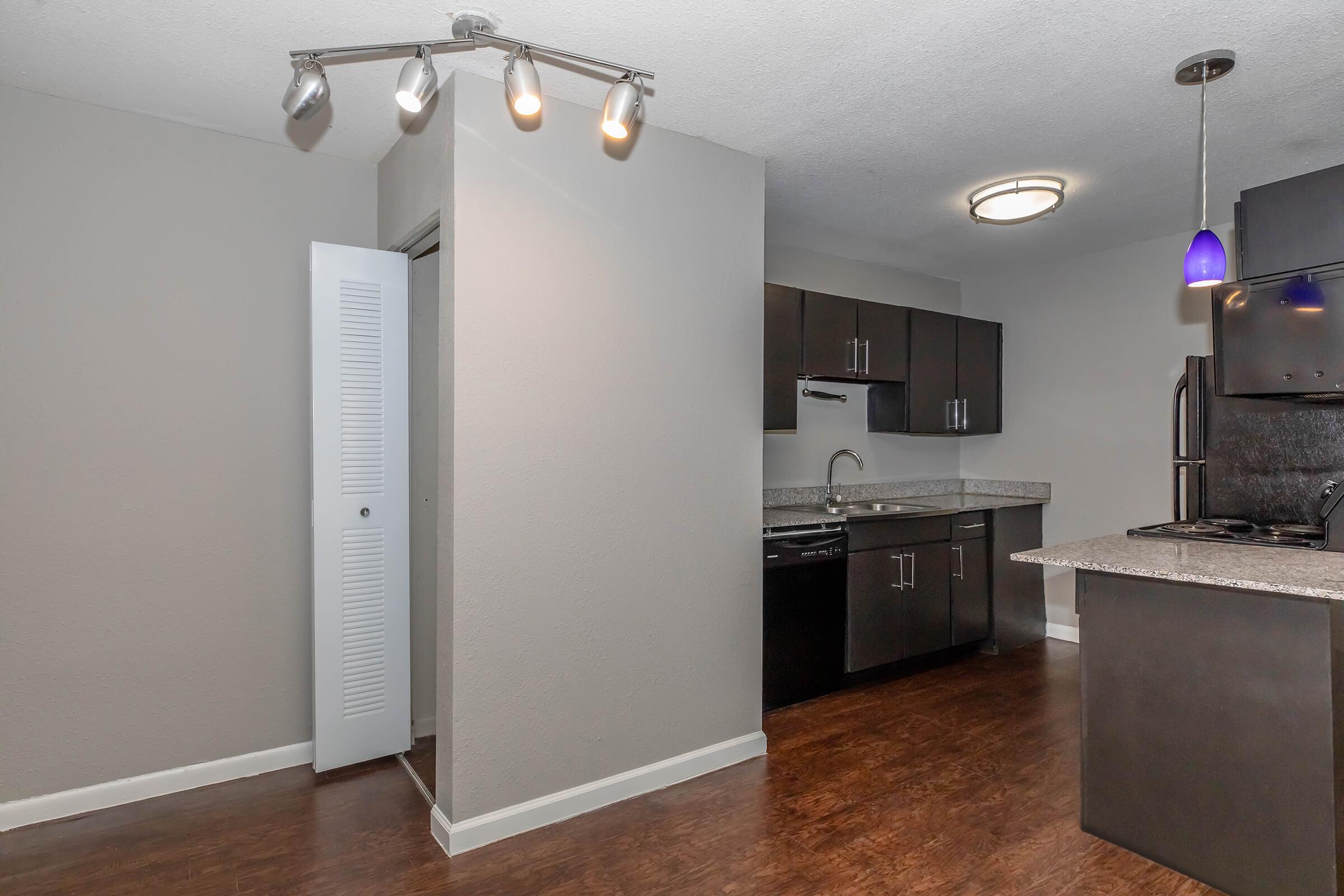 A modern kitchen with dark cabinetry and granite countertops. There is a stainless steel sink, refrigerator, and a stove, along with overhead lighting. The flooring is dark wood, and a doorway leads to another room. A pendant light hangs above the kitchen island. The walls are painted in a neutral color.