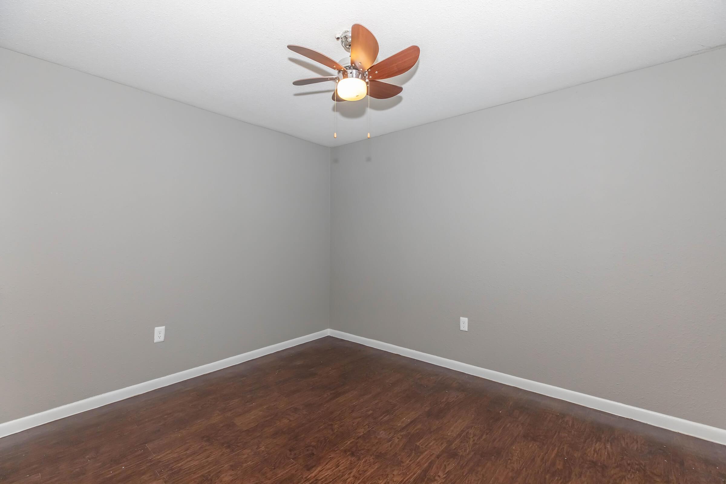 Empty room with light brown wooden flooring, gray walls, and a ceiling fan with wooden blades. The space is well-lit, featuring a minimalistic design with no furniture or decorations.