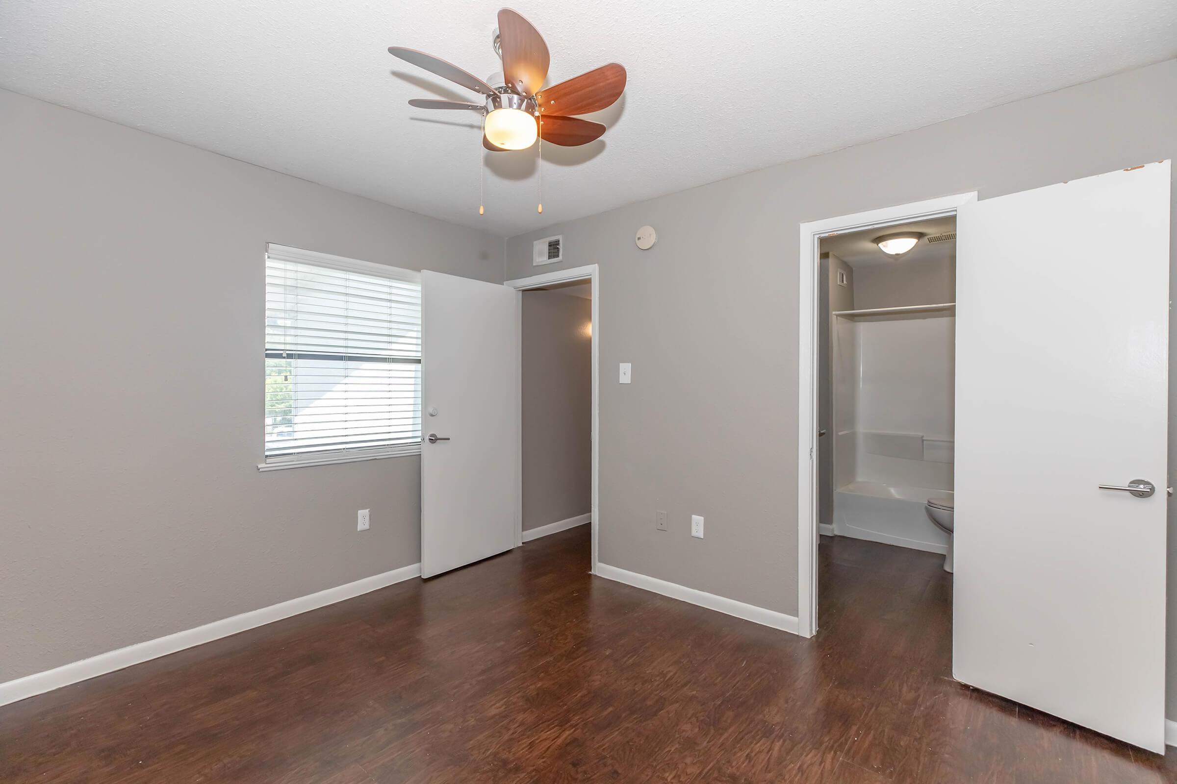 A well-lit room with light gray walls, featuring a ceiling fan with wooden blades. There are two white doors on the left side, one leading to another room and the other possibly to a closet. A small bathroom is visible through the door on the right, and the floor has dark wood-like laminate.
