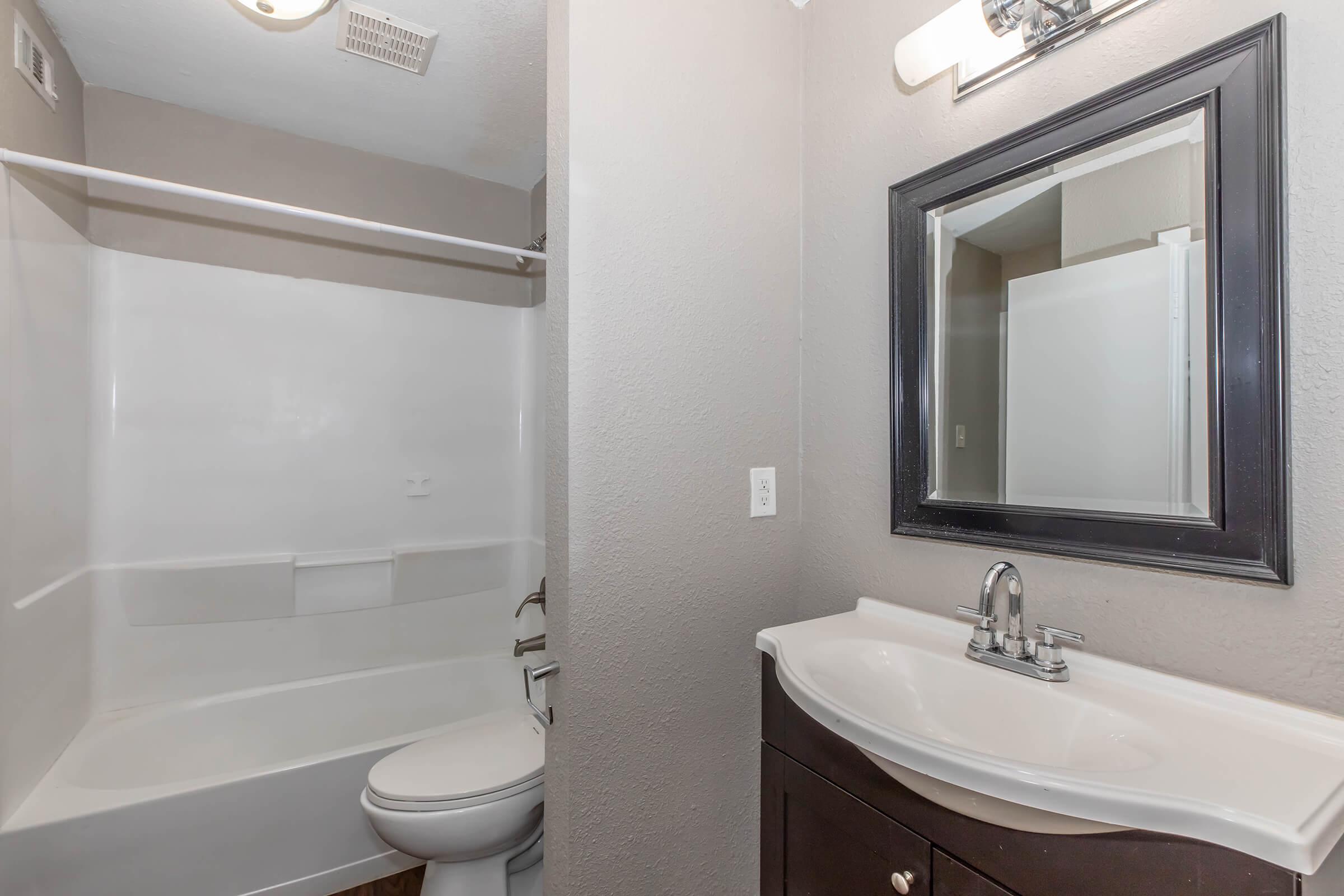 A small bathroom featuring a white bathtub with a shower, a modern black-framed mirror above a sleek sink with a faucet, and dark cabinetry. The walls are painted a light gray, and there is a light fixture on the ceiling. The space appears clean and functional.