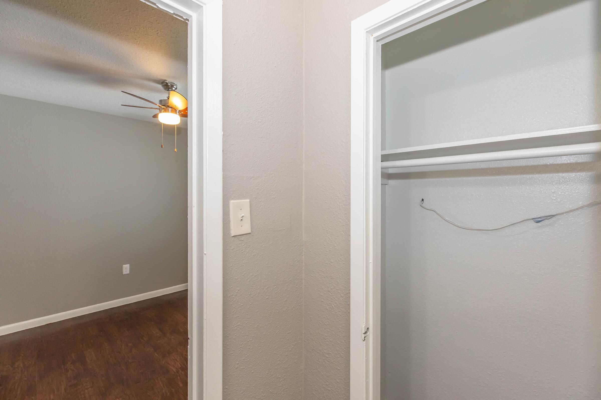 A view from a doorway leading to a closet on the left, featuring a single shelf and hanging space. The wall is painted gray, and a ceiling fan is visible in the room beyond the doorway. The floor has a wood-like appearance, contributing to a modern and spacious feel in the area.