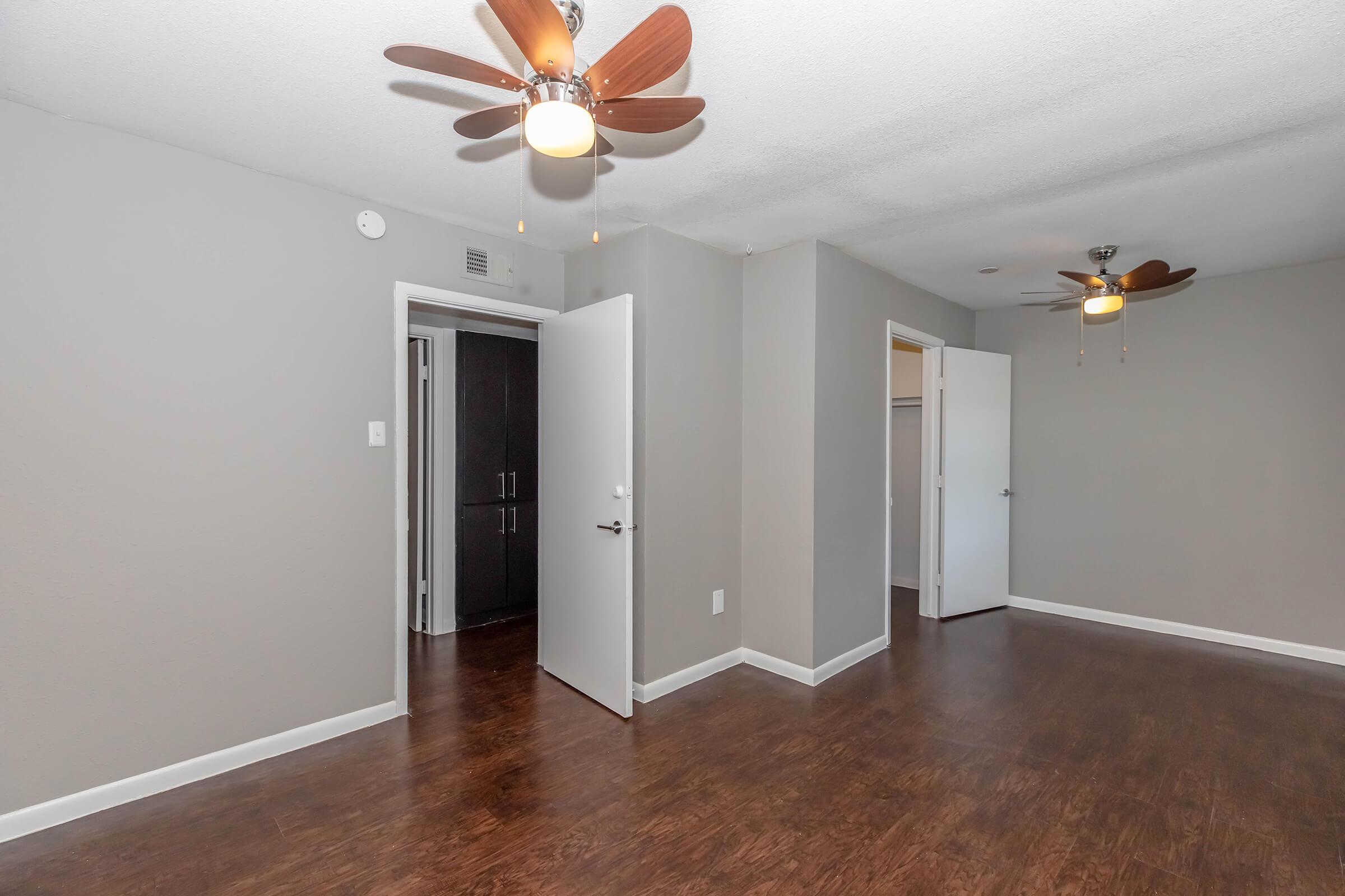 Interior view of a bedroom with two ceiling fans and modern light fixtures. The walls are painted light gray, and there are doors leading to a closet and another room. The floor is dark wood, creating a warm atmosphere. The room is well-lit and spacious, suitable for various furniture arrangements.