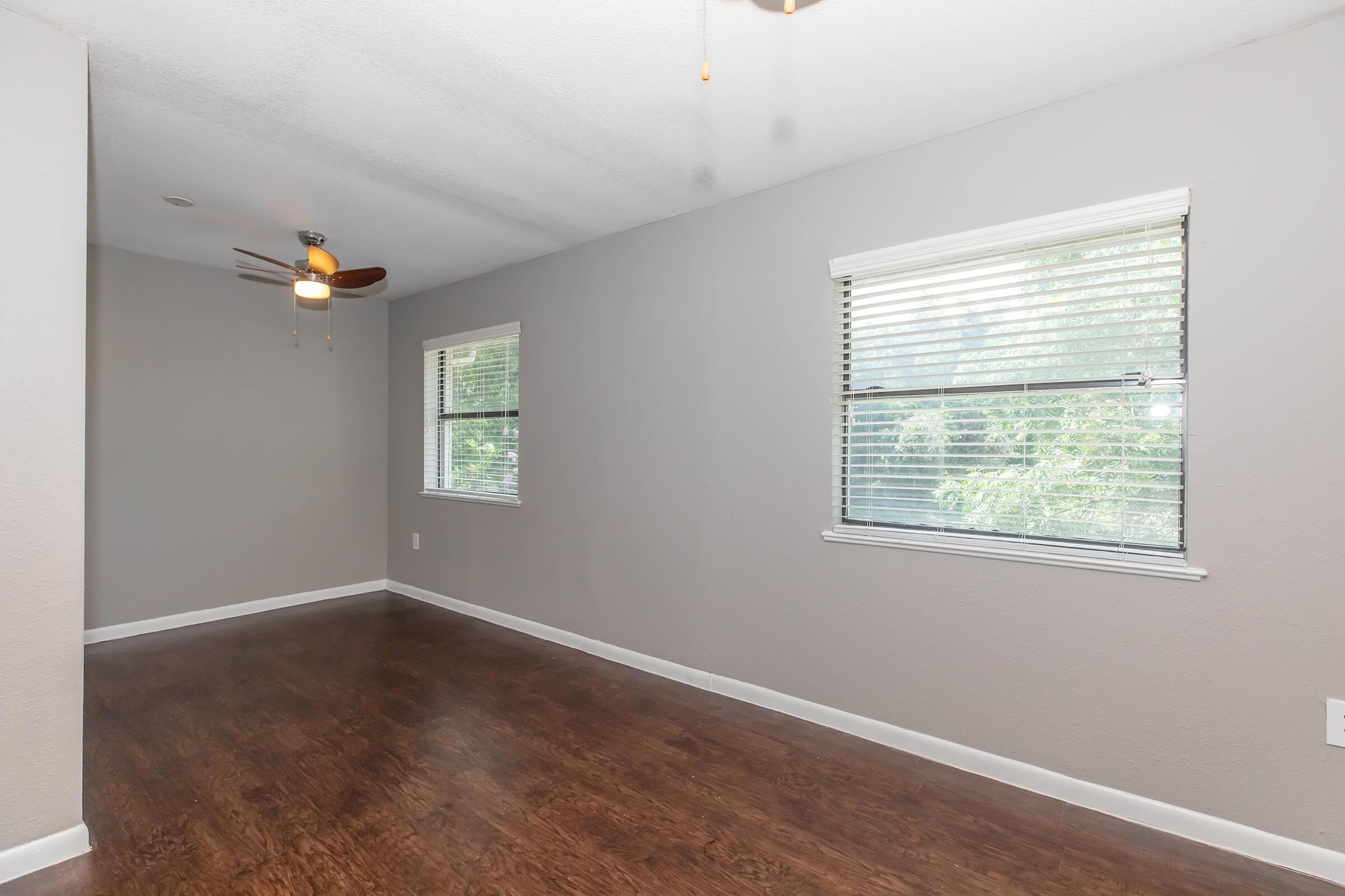 An empty room with light gray walls and a dark wood floor. It features two windows with white blinds on the left side, allowing natural light to enter. A ceiling fan is visible in the upper left corner. The overall space is bright and clean, suitable for various setups.