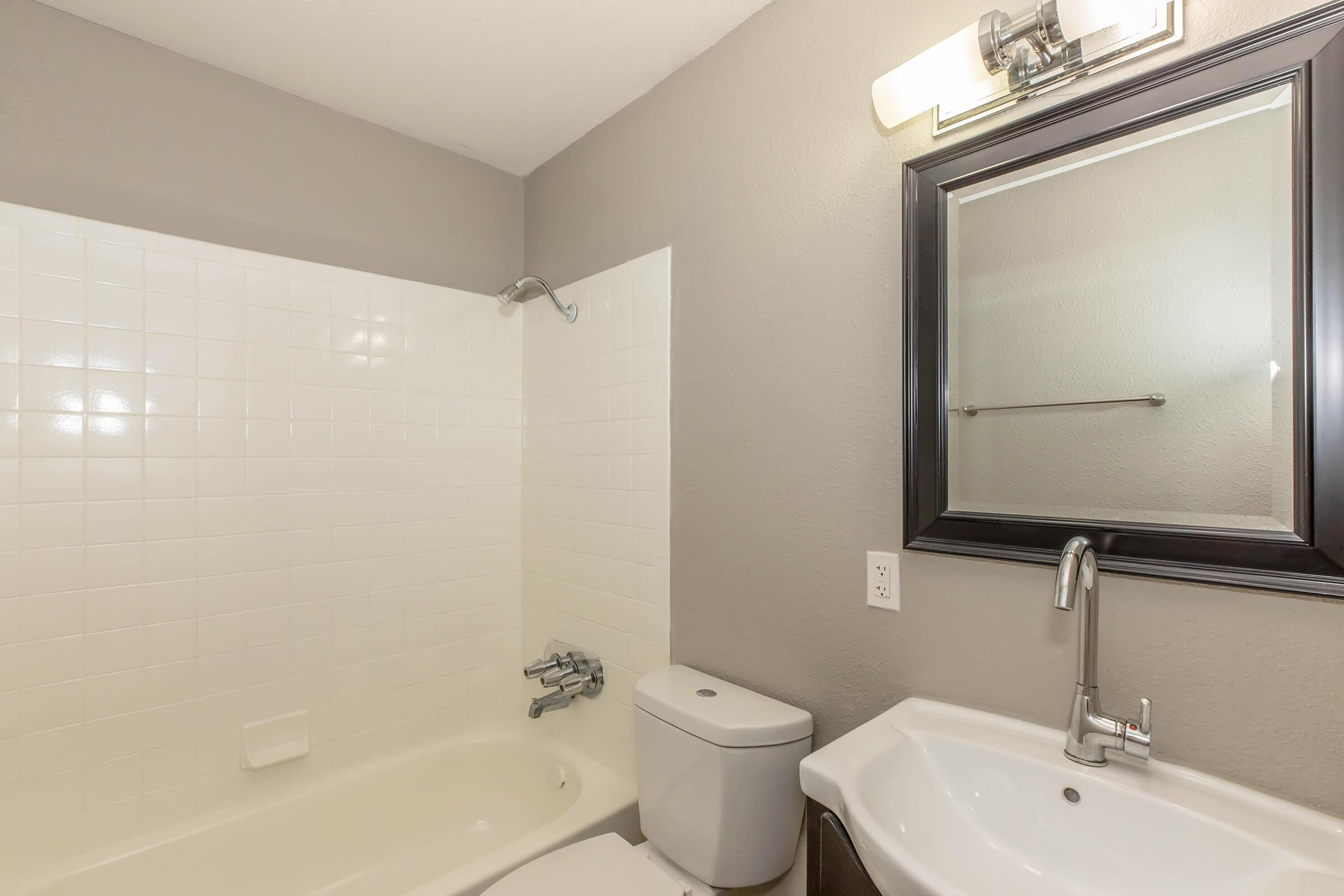 A modern, neutral-colored bathroom featuring a bathtub with a showerhead, a white toilet, and a sink with a sleek faucet. The walls are painted gray, and there is a rectangular mirror above the sink with a light fixture above it. The overall design is clean and minimalist.