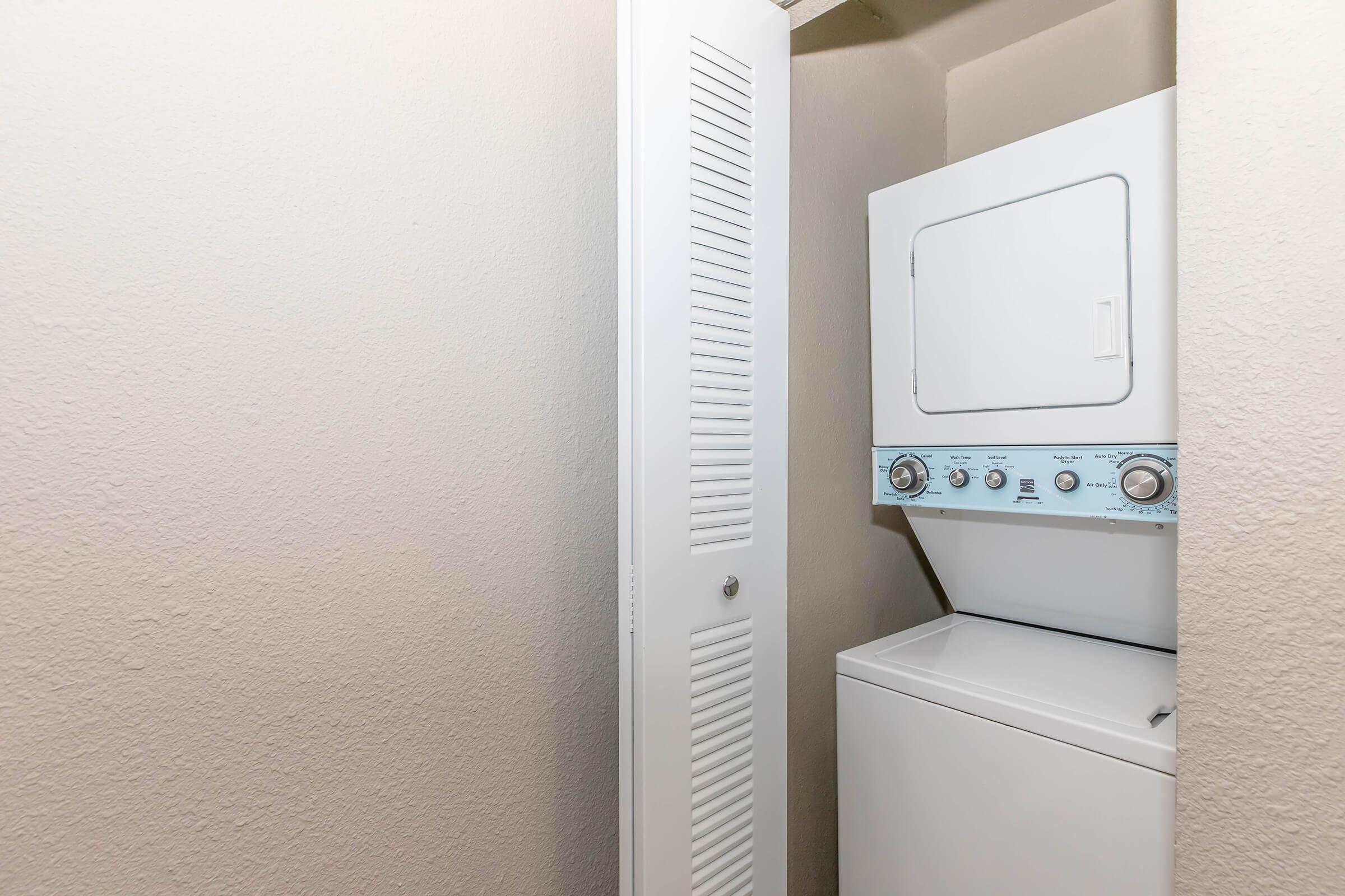 A stacked washer and dryer set in a small laundry closet. The walls are beige and there is a closet door beside the appliances. The washer is at the bottom, and the dryer is on top, with control knobs and a dial visible on the front.