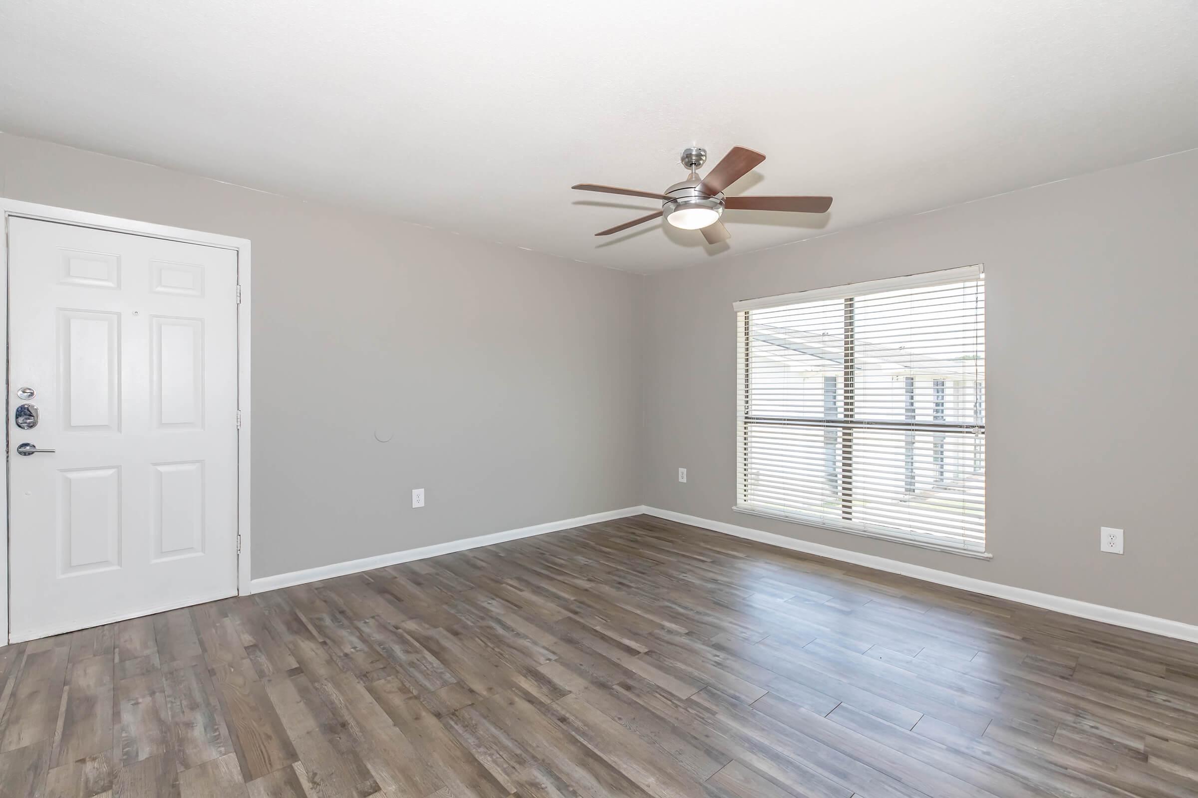 A spacious room with light gray walls and a brown wooden floor. A ceiling fan is mounted in the center, and there is a window with blinds that lets in natural light. An entrance door is visible to the left of the image.
