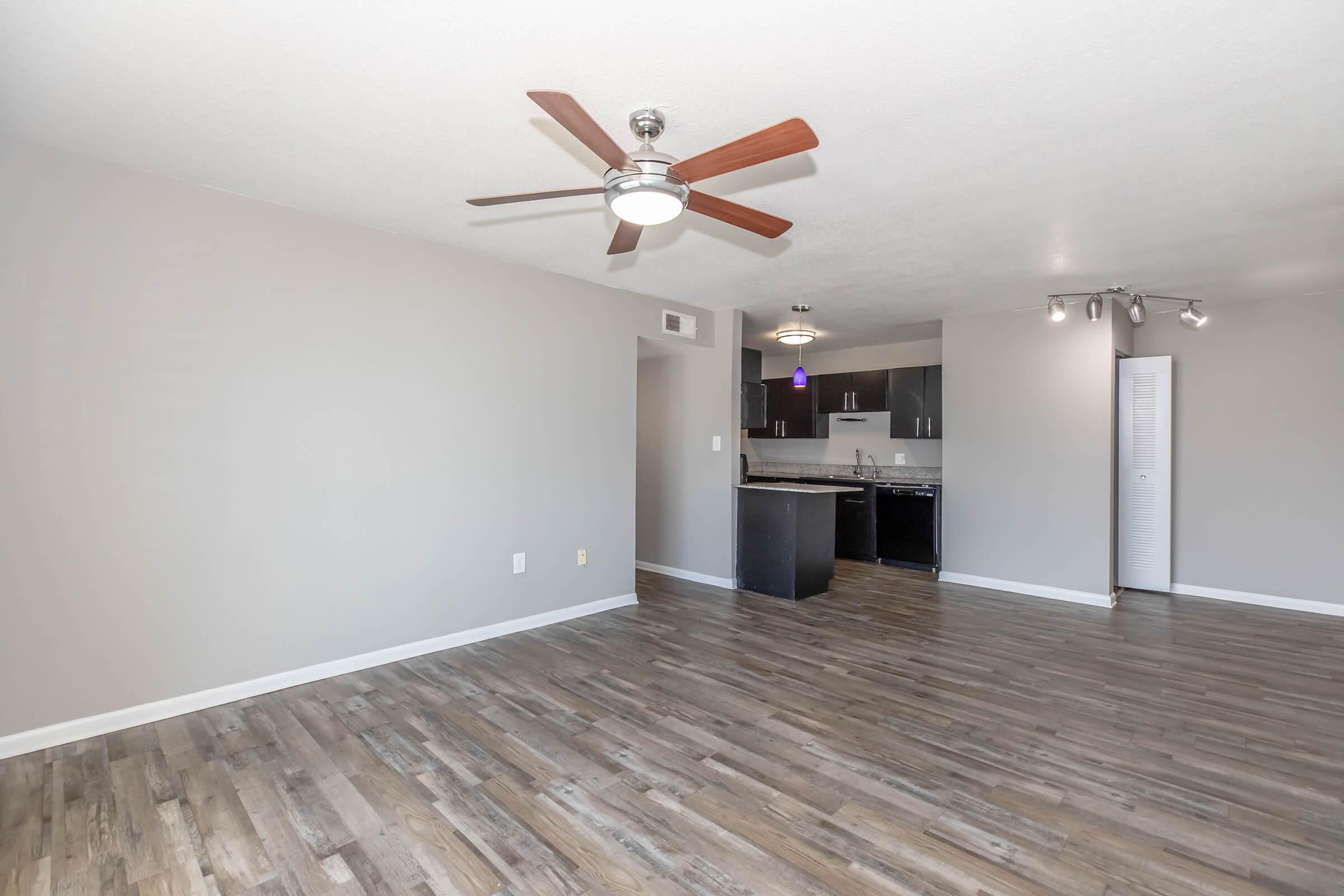 Spacious living area with wood-like flooring, a ceiling fan, and modern lighting. The open layout connects to a kitchen featuring dark cabinets and stainless steel appliances. Brightly lit with neutral-colored walls and a large entryway.