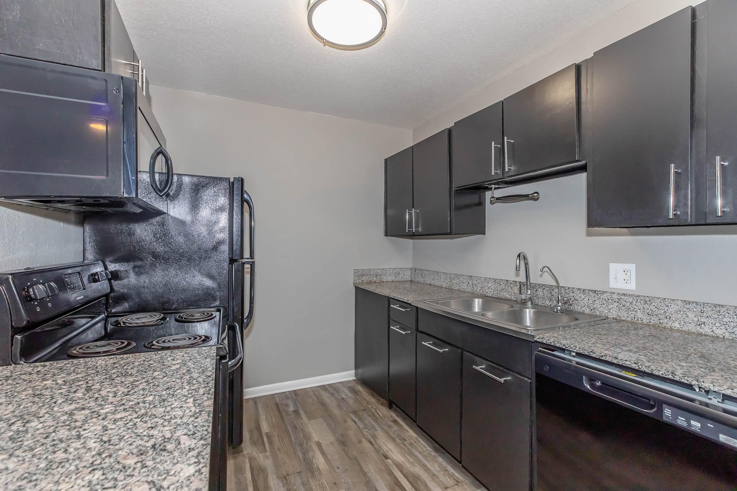 A modern kitchen featuring black cabinets and appliances, including a stove, refrigerator, and dishwasher. The countertops are made of granite, and there is a double sink with a modern faucet. The walls are painted in a light color, creating a bright and spacious atmosphere. The flooring is laminate wood.