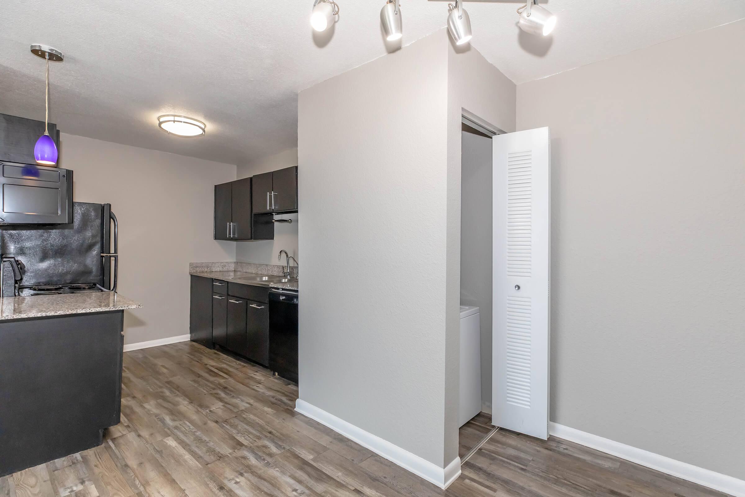 A modern kitchen and living area featuring dark cabinets, a granite countertop, and stainless steel appliances. The floor is made of wood-like tiles. A small laundry area is visible through an open door, and lighting fixtures hang from the ceiling, providing bright illumination.