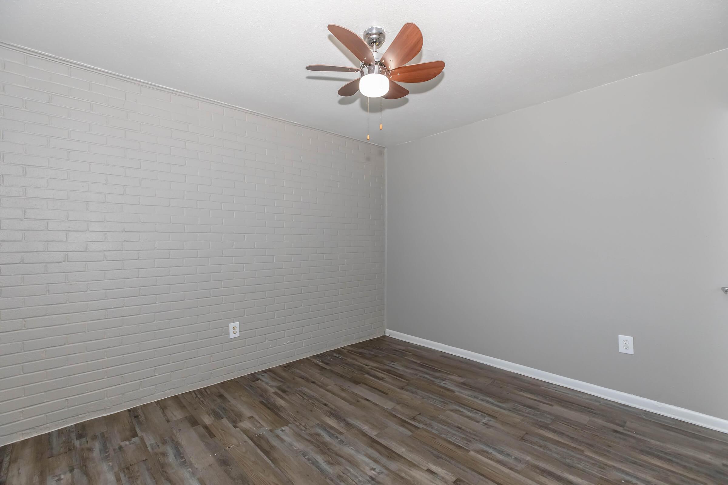 Empty room with a light gray brick accent wall, hardwood-style flooring, and a ceiling fan with wooden blades. The space has no furniture, creating a spacious and minimalistic look. Natural light may come from a window, adding brightness to the area.