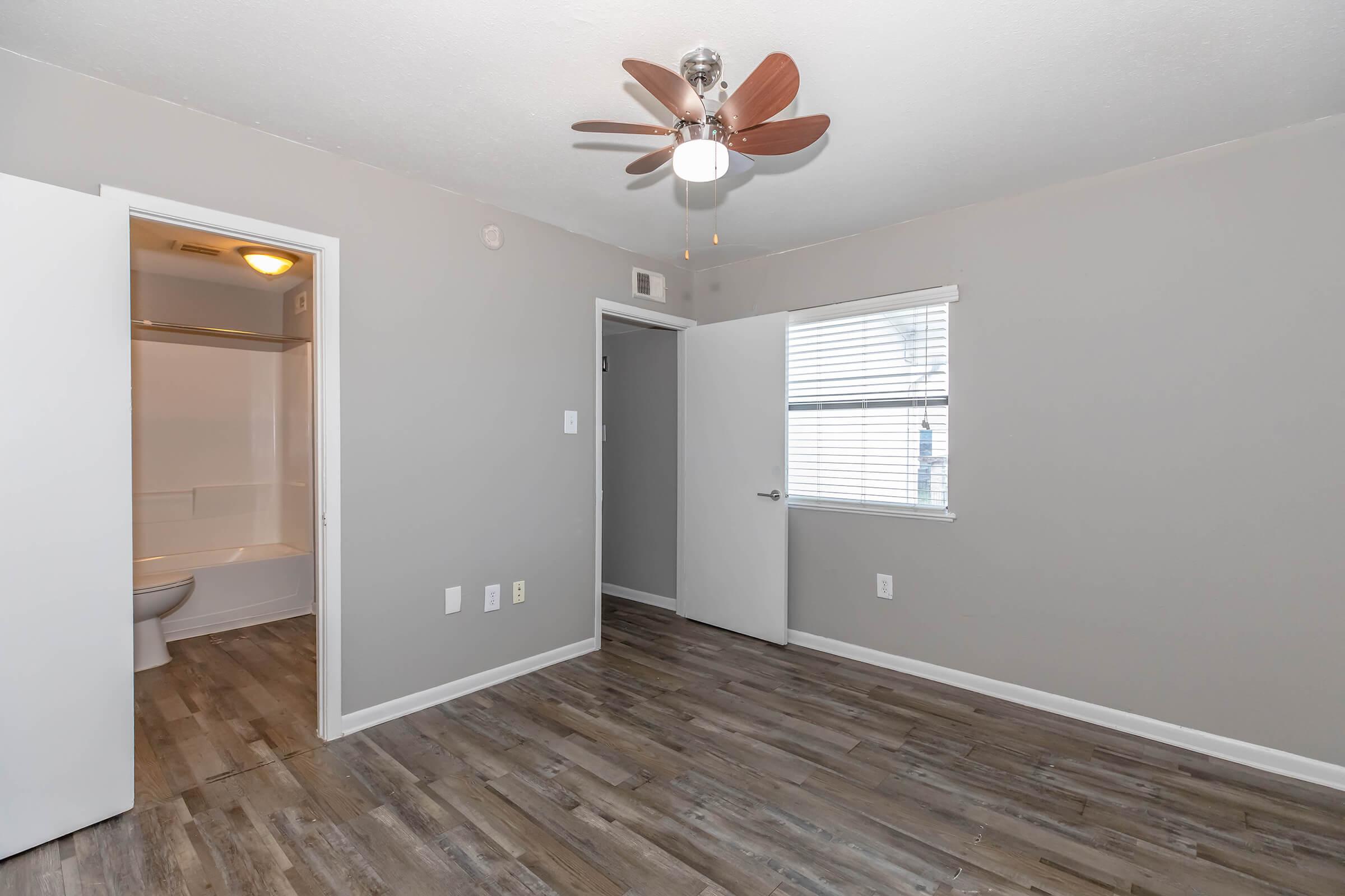 A bright, empty bedroom featuring gray walls and wood-style flooring. One wall has a window with blinds, allowing natural light in. A ceiling fan is centered above, and a door on the left leads to a bathroom. The room is clean, inviting, and ready for decoration.