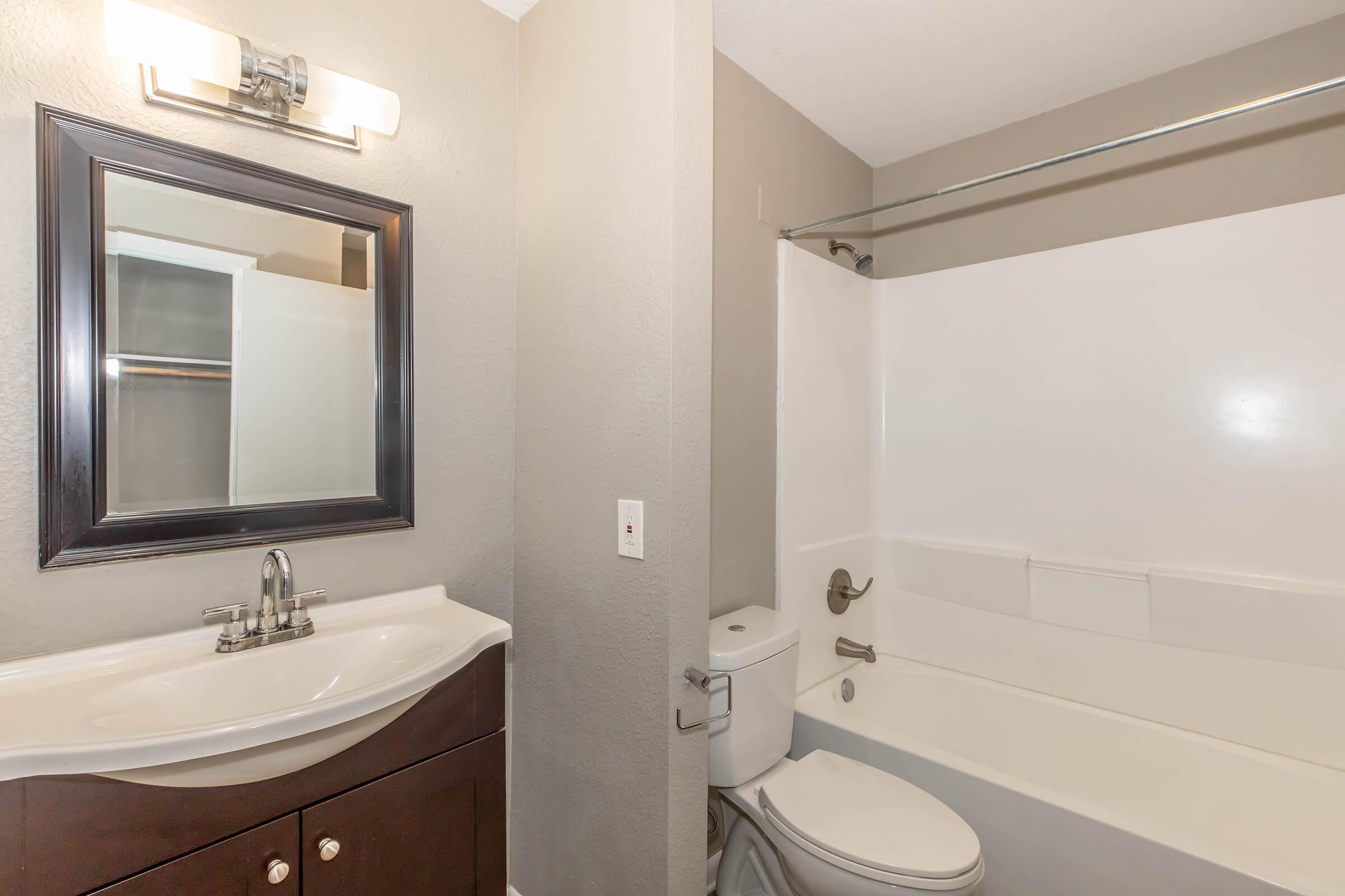 A modern bathroom featuring a white bathtub with a glass shower curtain, a sleek dark wood vanity with a sink, and a large square mirror above the vanity. Light-colored walls and a bright light fixture create a clean, neutral environment.