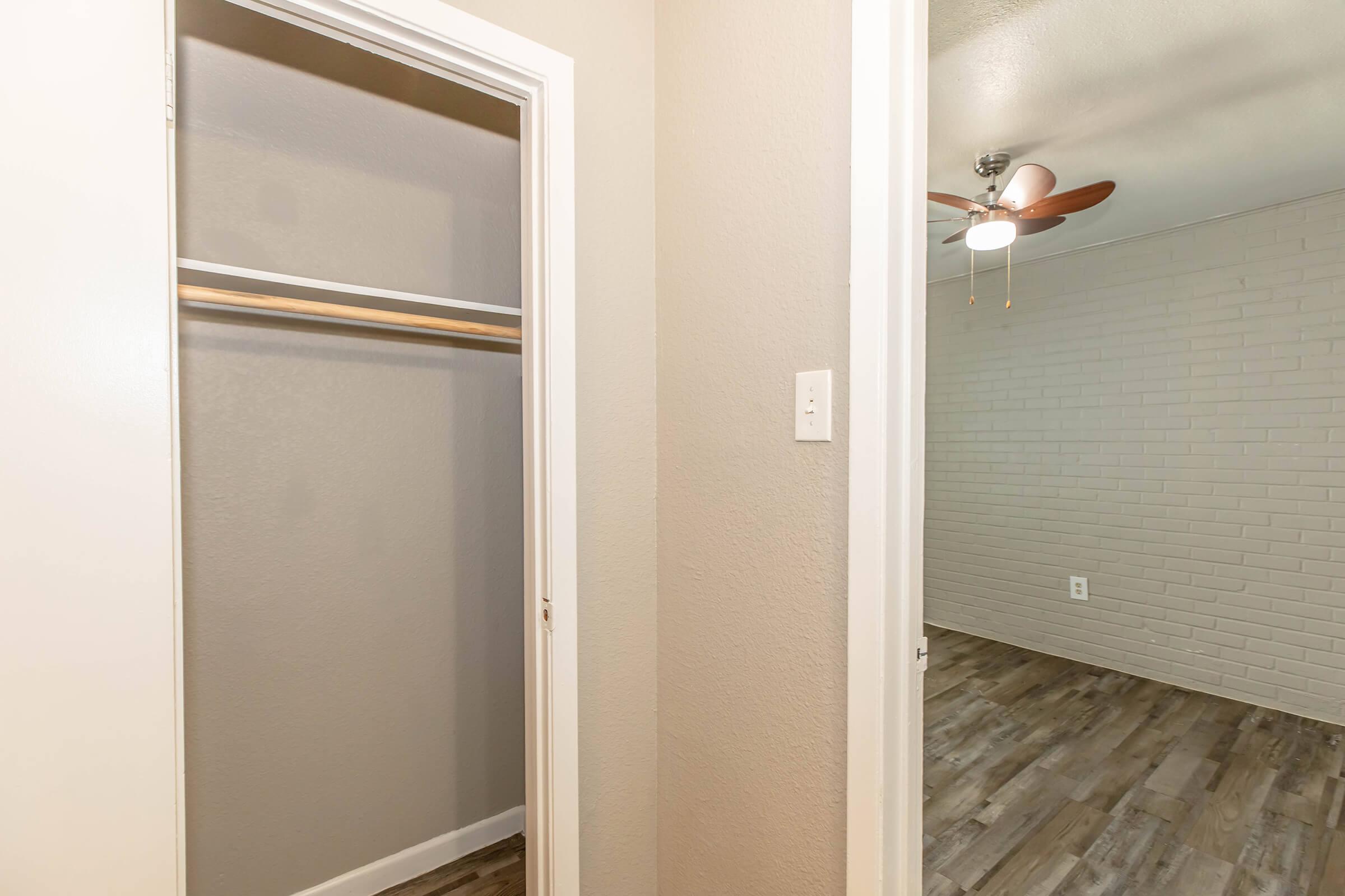 A doorway leading into a room with a light-colored wall featuring a ceiling fan. On the left, there's an open closet with a wooden rod for hanging clothes. The floor is covered in wood-like laminate, and the room appears well-lit and spacious.