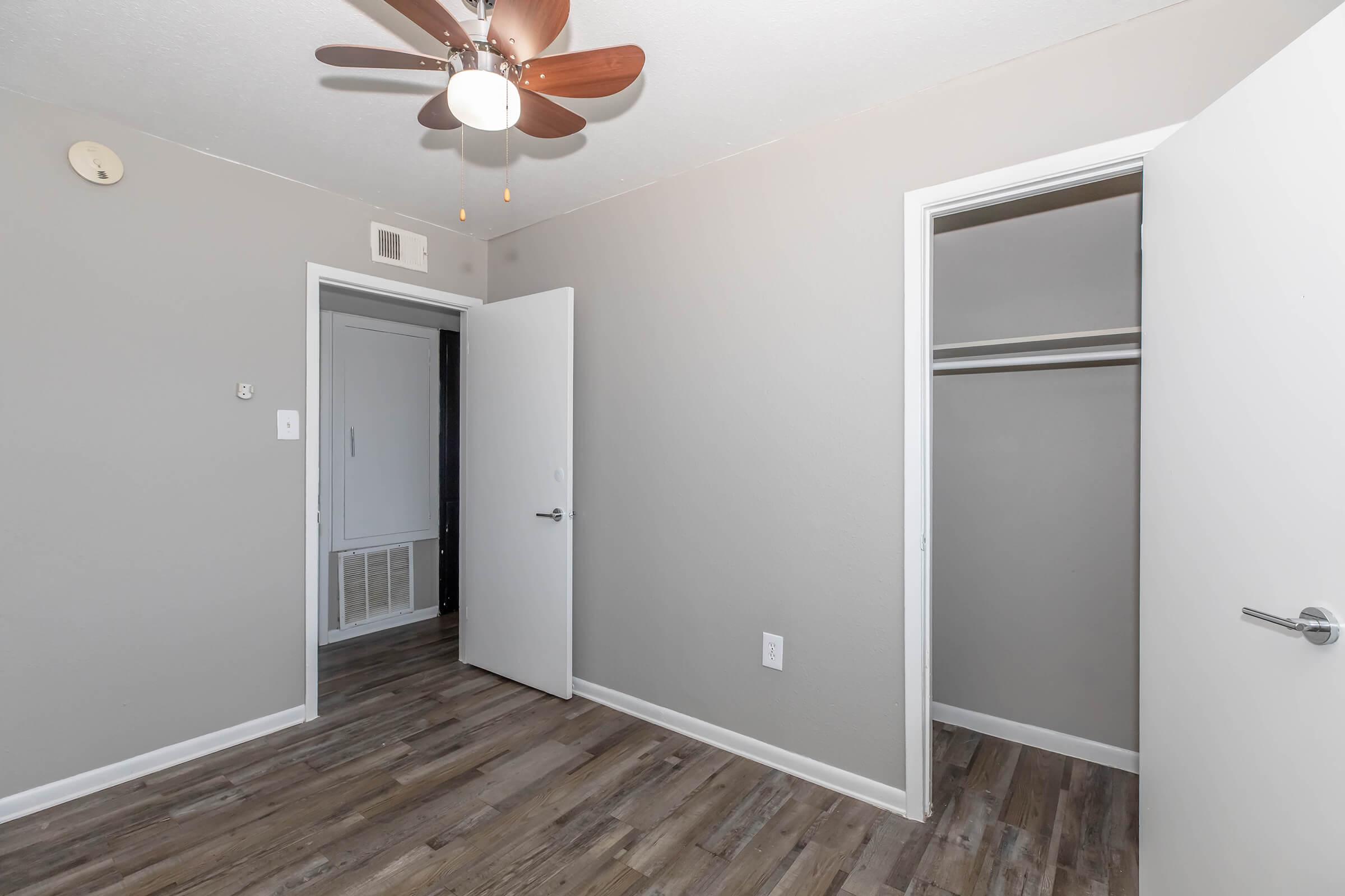 A well-lit empty room featuring a ceiling fan, two white doors leading to a closet and another room. The walls are painted in a light gray color, and the floor has wood-like laminate. There is a small vent on the wall and a light fixture on the ceiling.