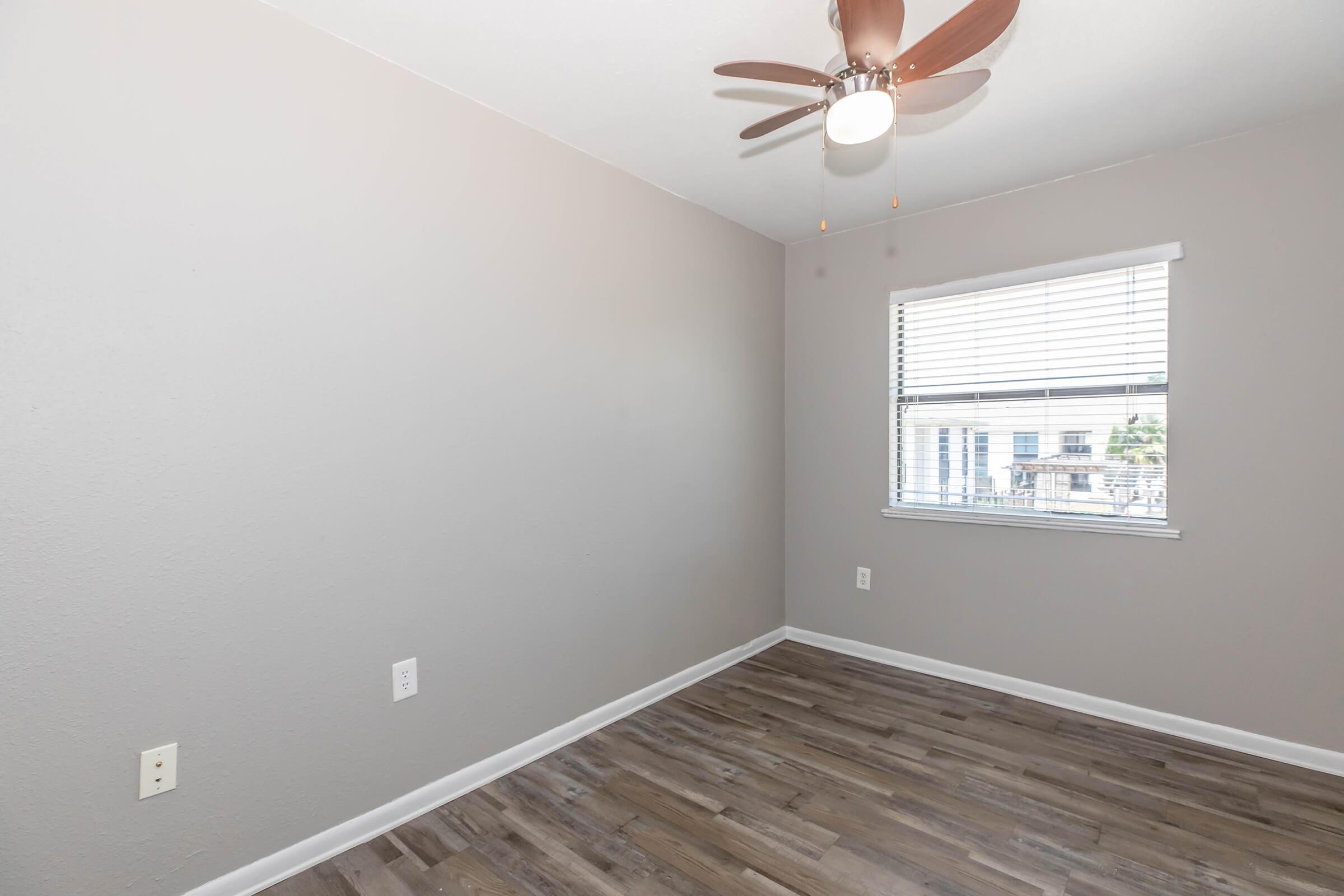 Empty room featuring gray walls and laminate flooring. A ceiling fan with wooden blades hangs above, and there's a window with white blinds letting in natural light. The room has minimal decor, providing a spacious and clean appearance.