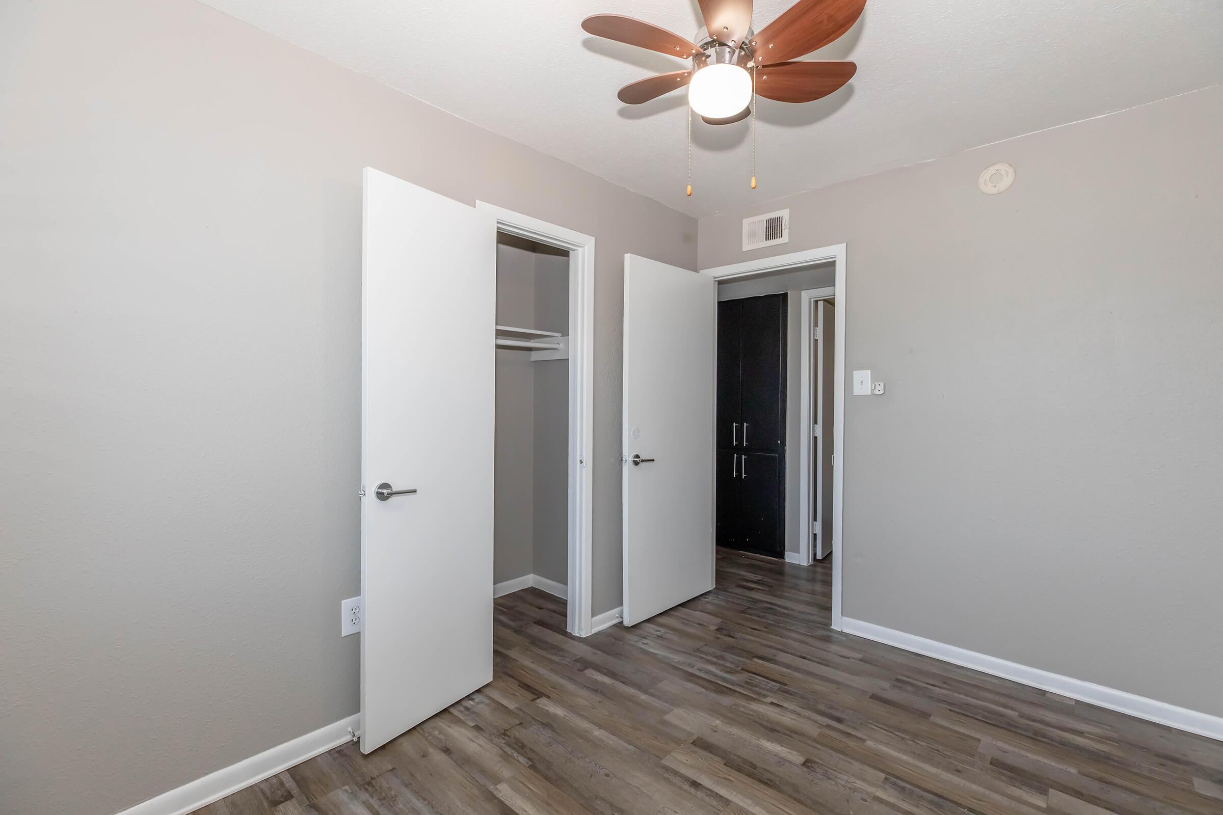 A spacious, lightly colored bedroom featuring a ceiling fan, two open white doors leading to a closet, and wooden floors. The walls are painted in a neutral tone, enhancing the brightness of the room. A dark door at the far end hints at another room or closet.