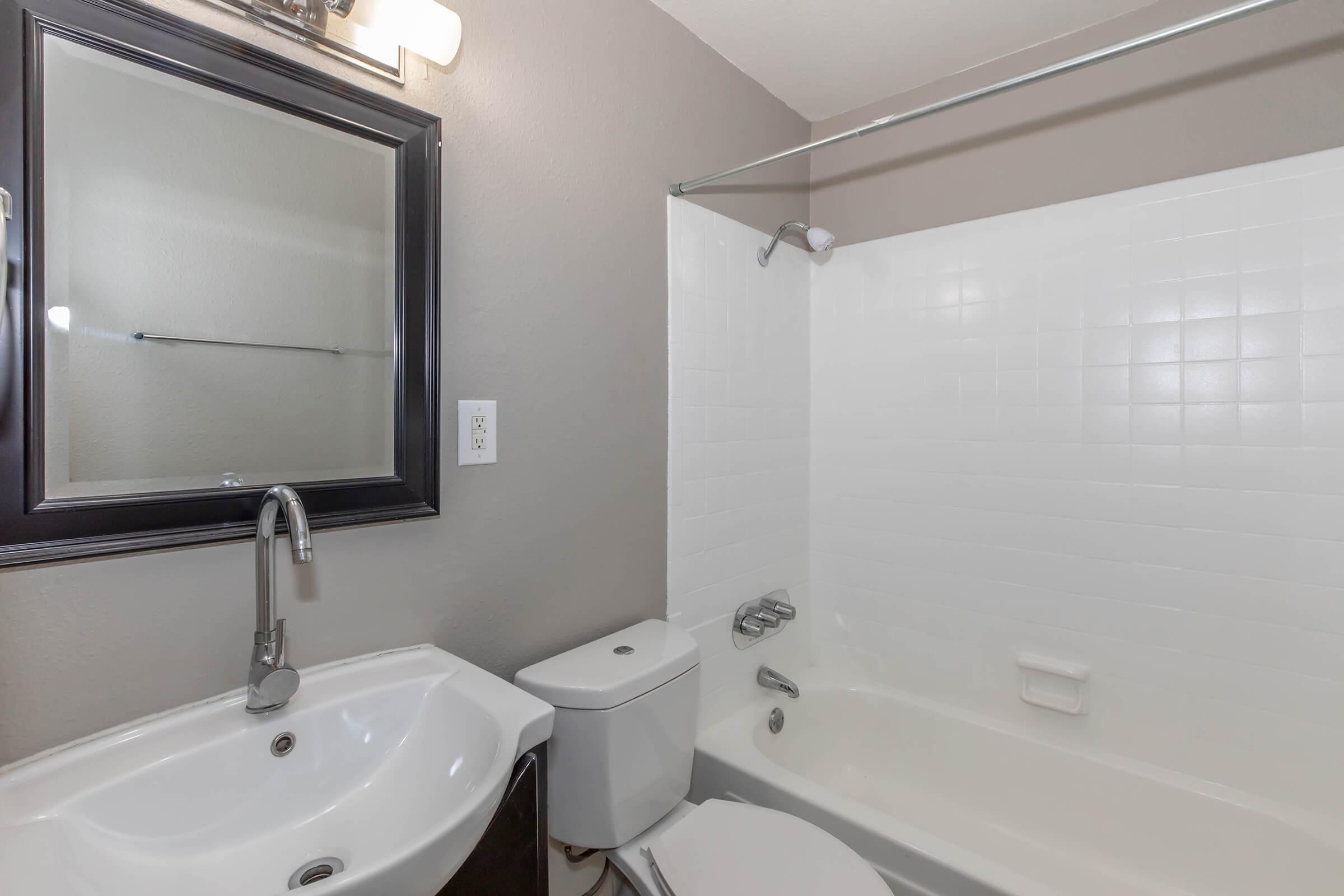 A modern bathroom featuring a white bathtub and shower, a wall-mounted mirror in a black frame, a sleek faucet above a contemporary sink, and a white toilet. The walls are painted a neutral gray color, adding a clean and minimalist aesthetic to the space.
