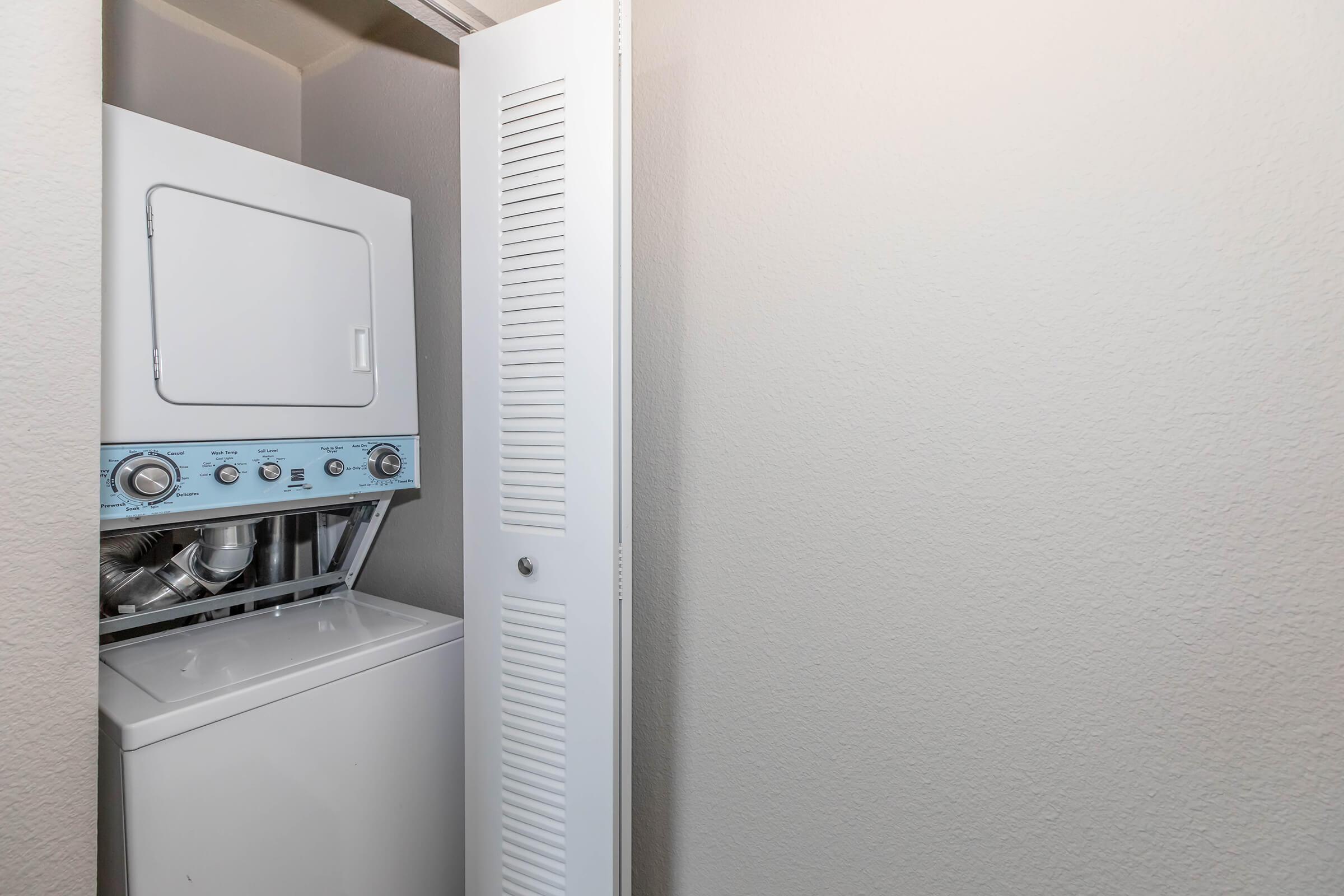 A compact laundry area featuring a stacked washer and dryer unit, located in a narrow closet space. The walls are painted in a light color, and a louvered door is partially visible on the right side, enhancing the organized appearance of the space.