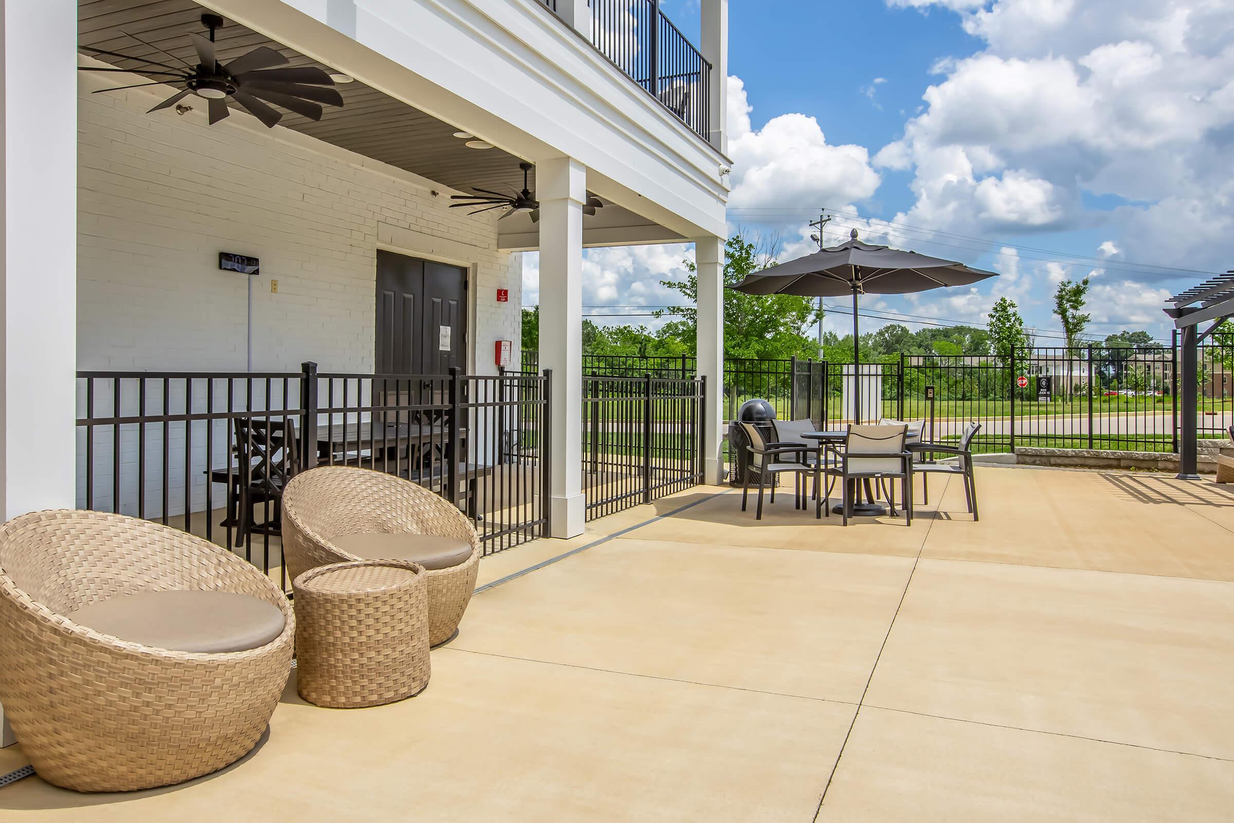 Outdoor patio area featuring two woven lounge chairs, a dining table with chairs, and an umbrella. The space is surrounded by a black fence, with trees and grass visible in the background under a partly cloudy sky.
