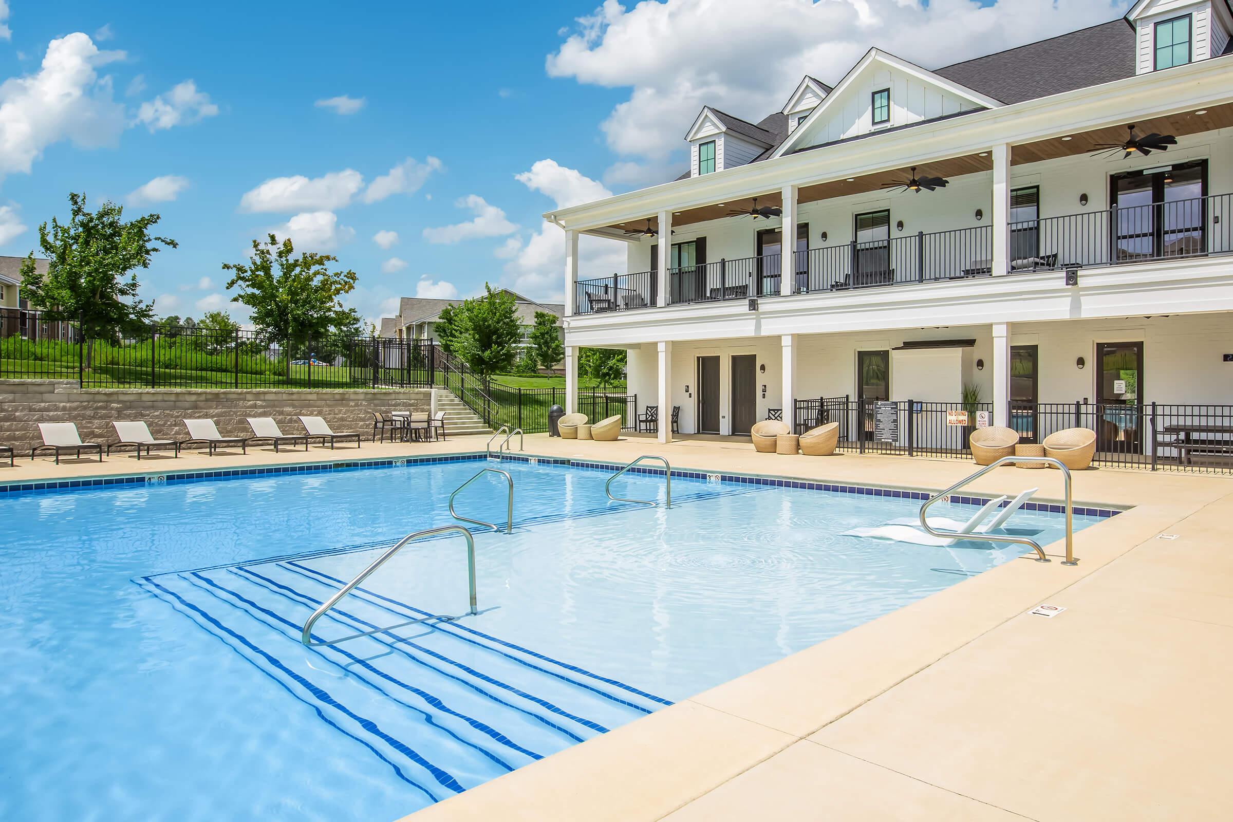 A scenic outdoor pool area featuring a large swimming pool with safety ladders, surrounded by lounge chairs and manicured landscaping. The pool is adjacent to a modern two-story building with balconies, large windows, and a clear blue sky with scattered clouds above.
