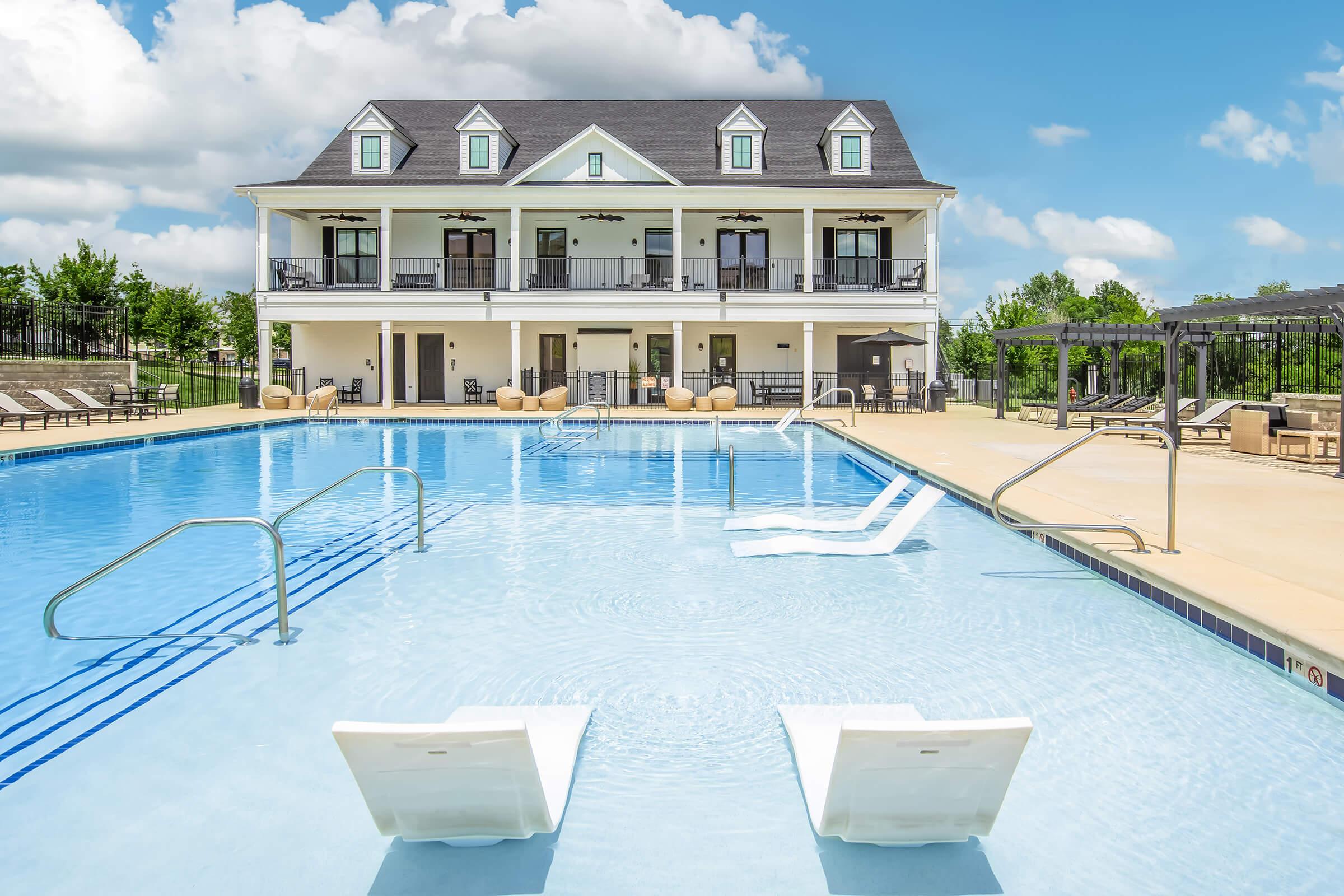 A clear blue swimming pool with lounge chairs partially submerged, surrounded by a well-maintained lawn. In the background, a modern two-story building features balconies and multiple entrance doors, under a bright blue sky with fluffy white clouds.