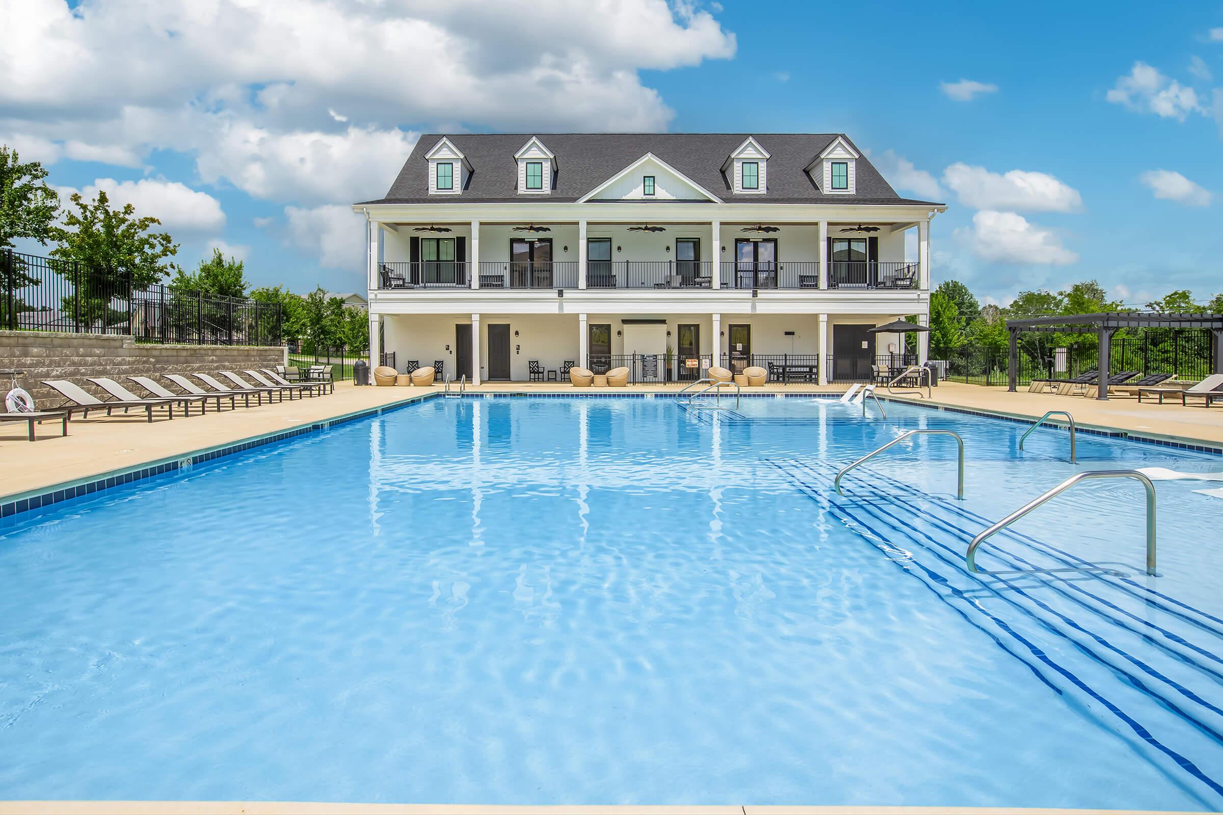 A clear blue swimming pool with a shallow area in the foreground, surrounded by lounge chairs. In the background, a two-story building features multiple balconies and large windows. The sky is partly cloudy, and landscaped greenery is visible around the pool area.