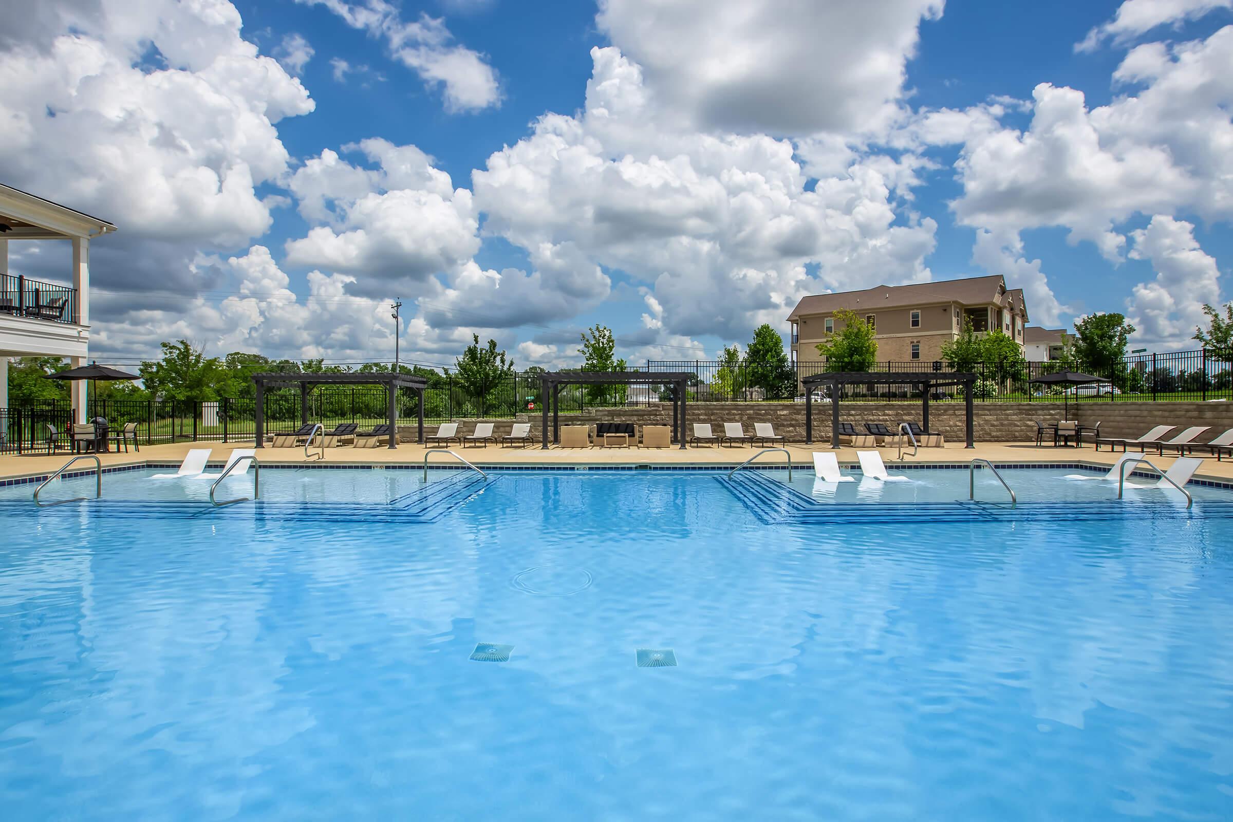 A bright blue swimming pool surrounded by lounge chairs and lush greenery, under a sky filled with fluffy white clouds. The pool features a shallow entry area and a spacious deck, creating a relaxing atmosphere for outdoor enjoyment.