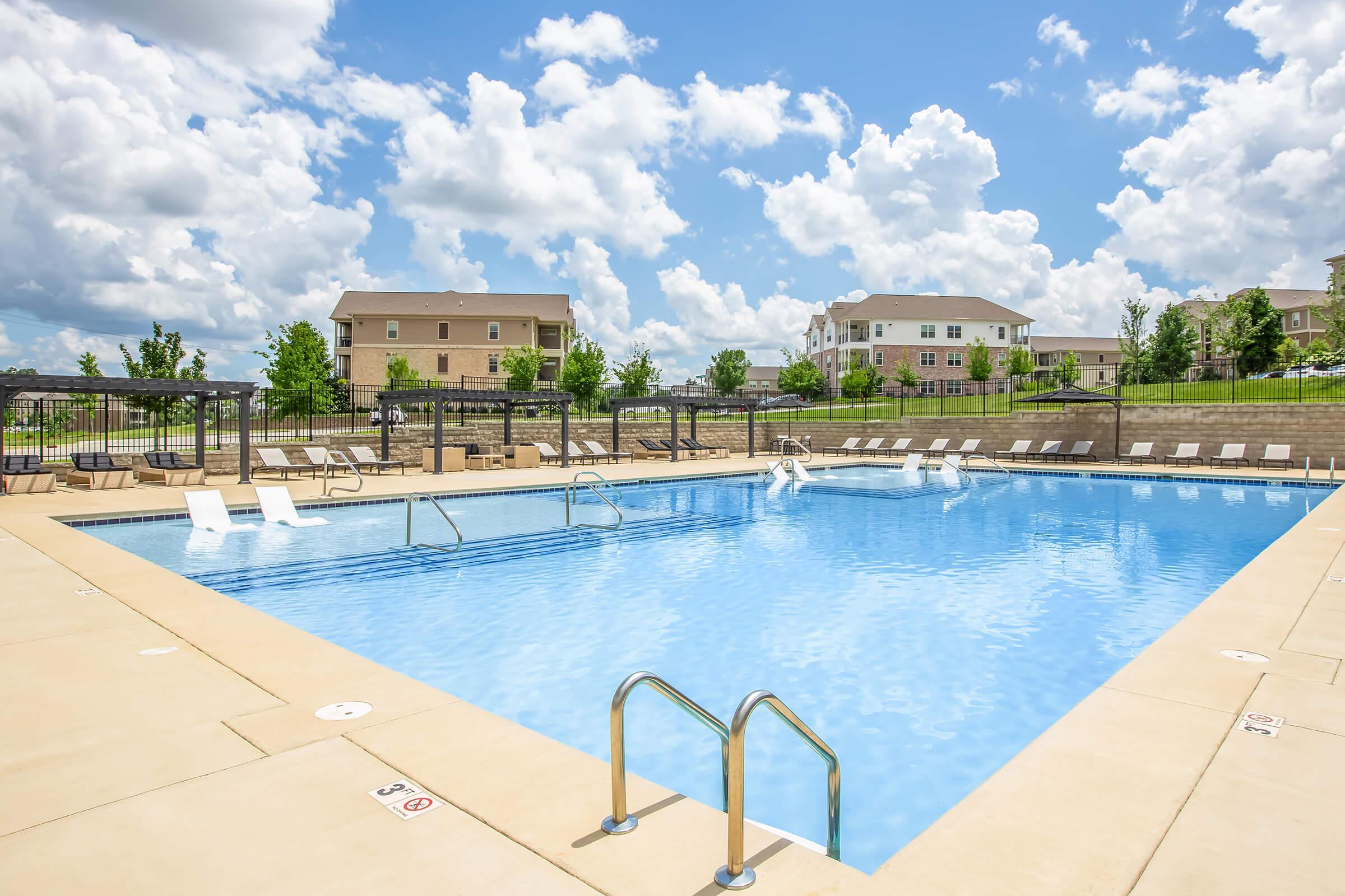 A spacious outdoor swimming pool surrounded by patio seating and lounge chairs, set against a backdrop of blue skies with fluffy clouds and green landscaping. Multiple lanes for swimming are visible, and there are residential buildings in the distance.