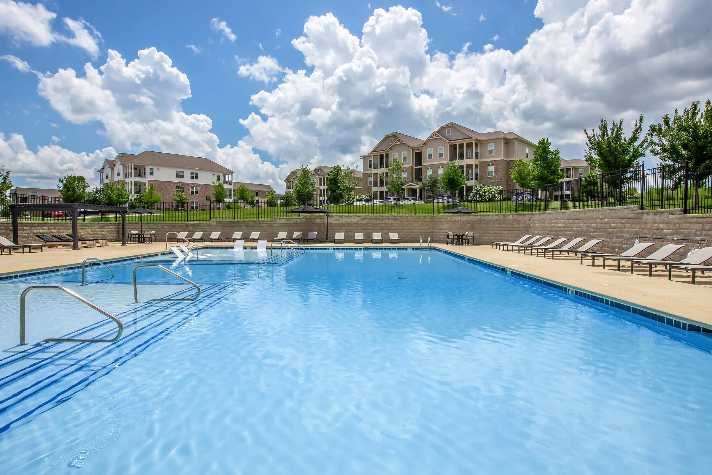 A clear blue swimming pool surrounded by lounge chairs and green trees, with several apartment buildings visible in the background. The sky is bright with fluffy clouds creating a vibrant, sunny atmosphere. The pool area features steps leading into the water, inviting relaxation and leisure.