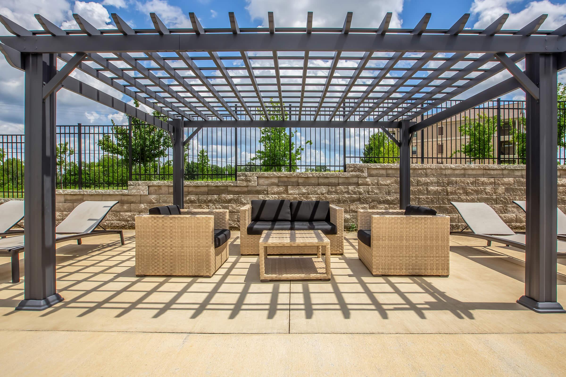 A cozy outdoor seating area featuring a pergola with a grid design overhead. The space includes two wicker chairs, a matching coffee table, and a backdrop of greenery and stone walls. Sun loungers are positioned nearby under a bright blue sky with scattered clouds.