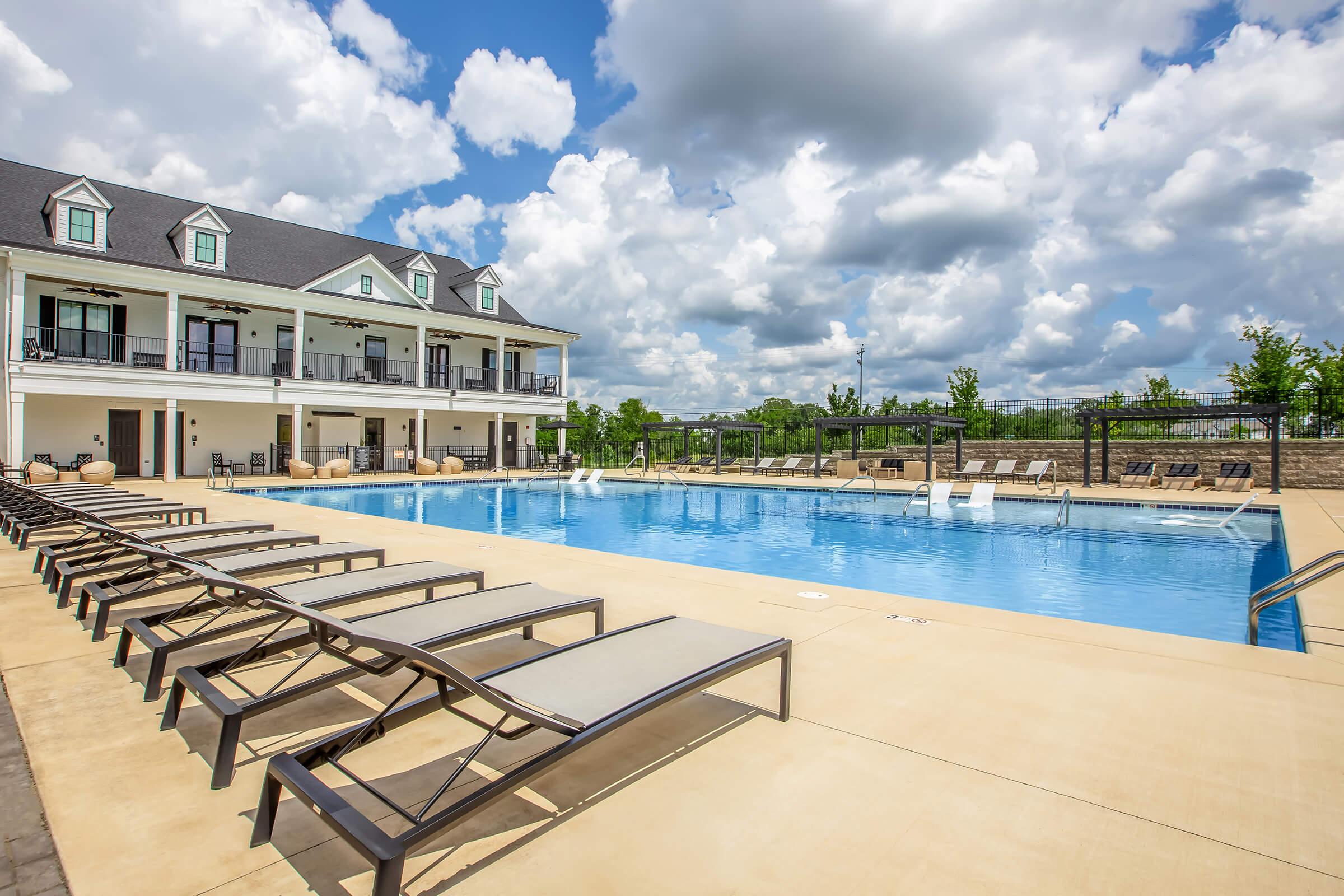 A clear blue swimming pool surrounded by lounge chairs, with a modern building in the background. The sky is partly cloudy, featuring large, fluffy white clouds. Green trees are visible in the distance, creating a serene outdoor setting.