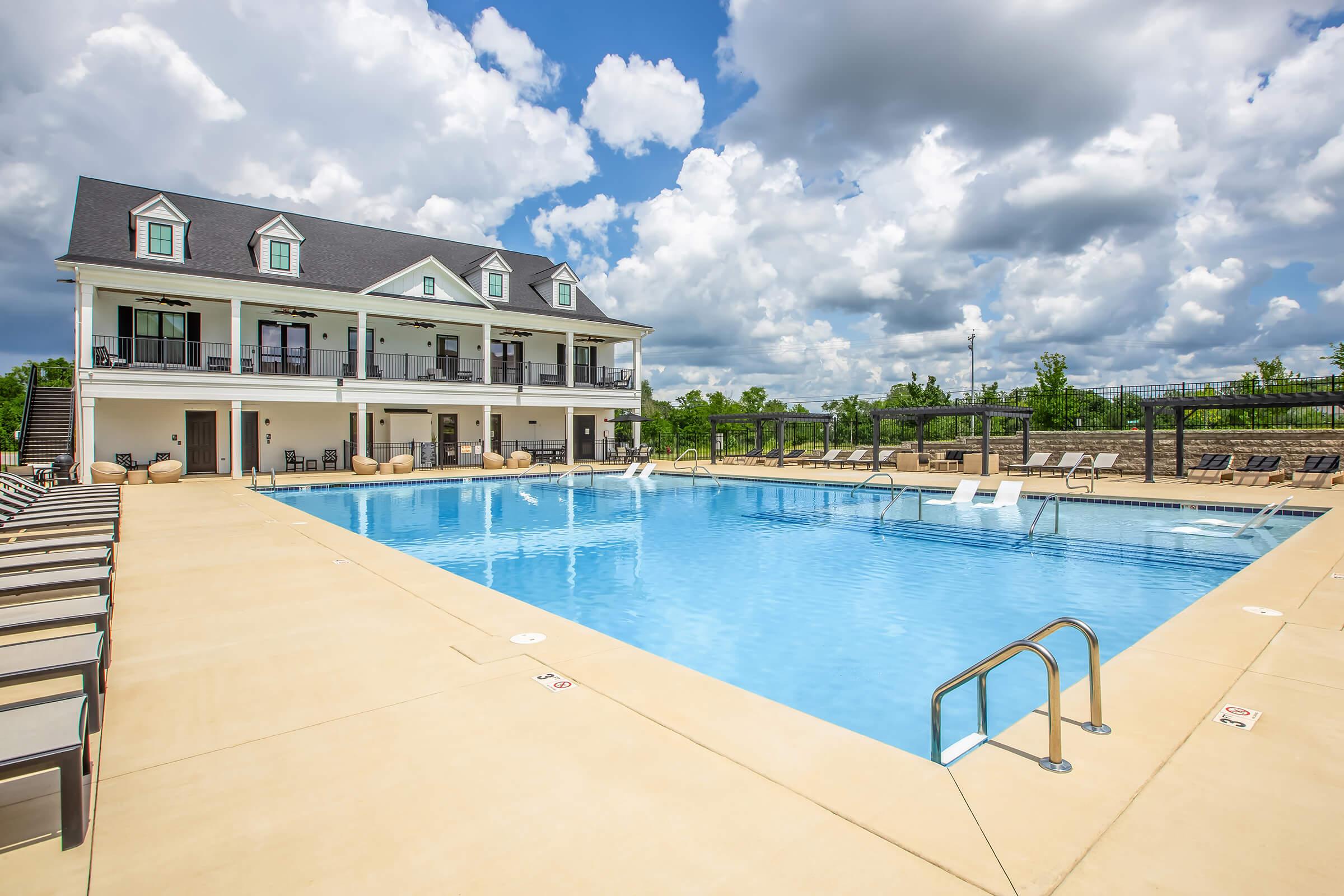 A spacious outdoor swimming pool surrounded by lounge chairs, with a two-story building in the background. The sky is partly cloudy, showcasing a bright day. The pool area features a clean, modern design with a relaxing atmosphere.