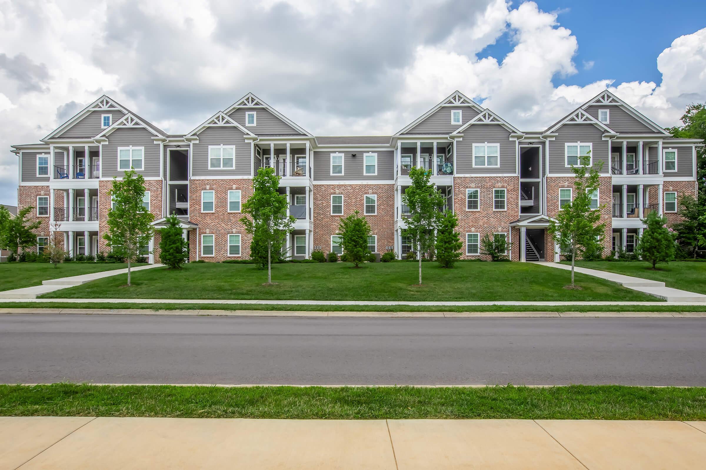 A row of modern, three-story apartment buildings made of brick and siding, featuring multiple balconies. The buildings are surrounded by lush green lawns and young trees, with a clear blue sky and fluffy clouds in the background. A sidewalk runs along the front, leading to a street.