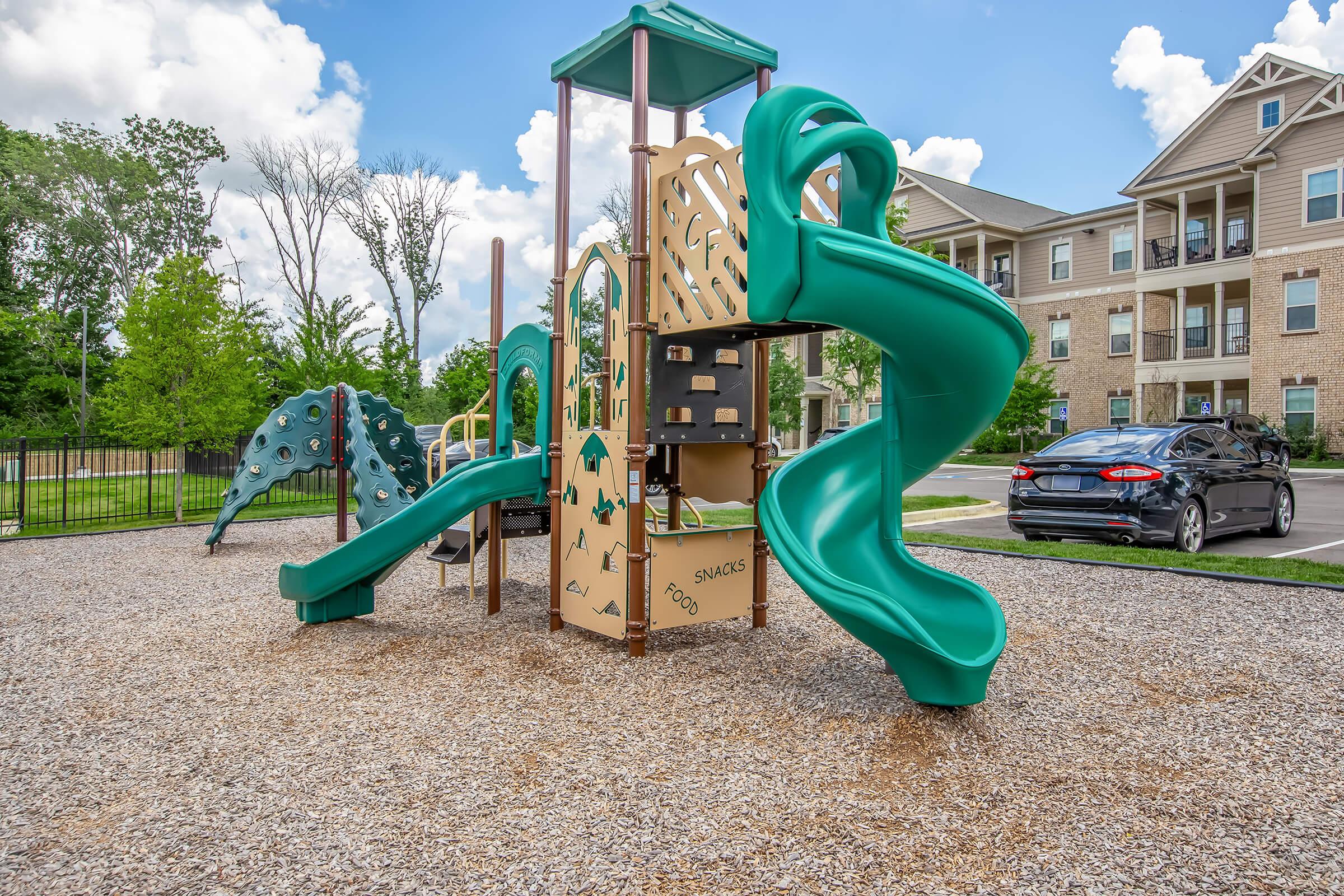 A colorful playground with green slides and climbing structures, set in a gravel area. In the background, there are trees and an apartment building. A parked car is visible nearby. The sky is blue with fluffy white clouds.