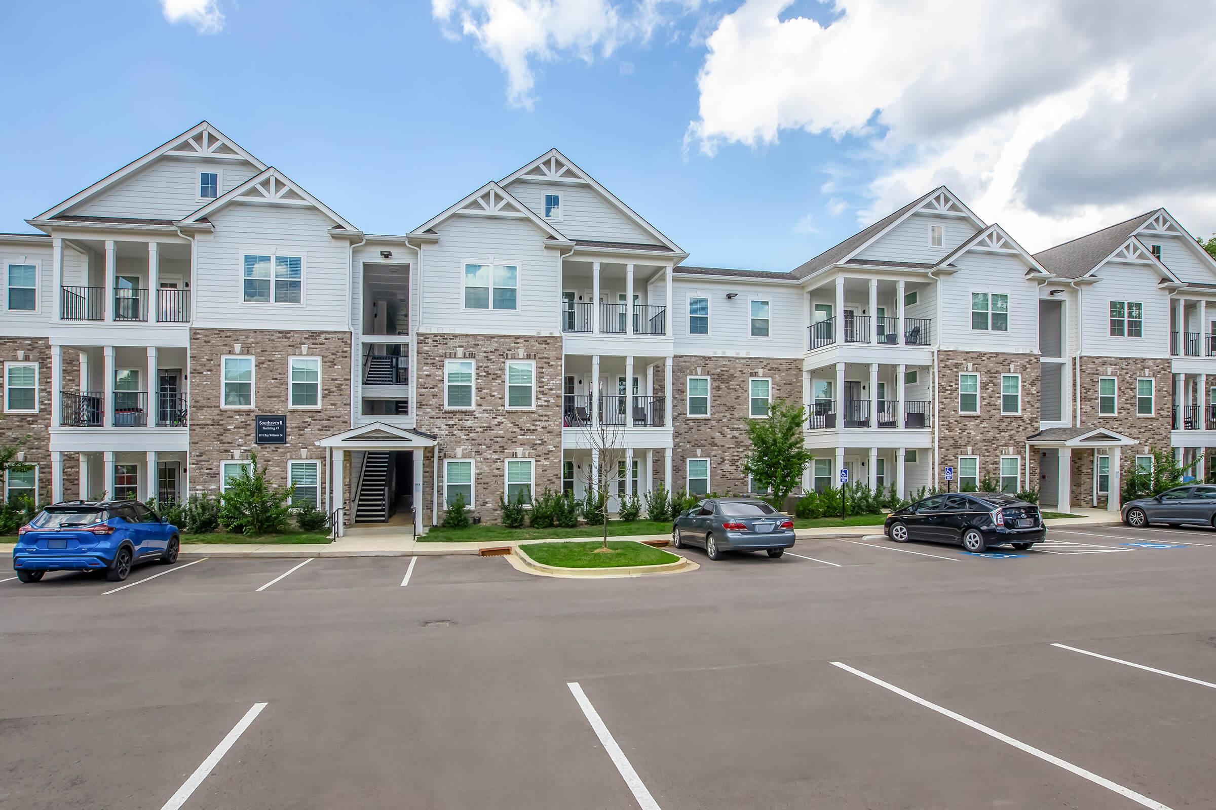 A modern apartment complex featuring multiple white and stone-finished buildings with balconies. The foreground shows a paved parking lot with several parked cars, and green landscaping surrounds the area. The sky is partly cloudy, indicating an overcast day.