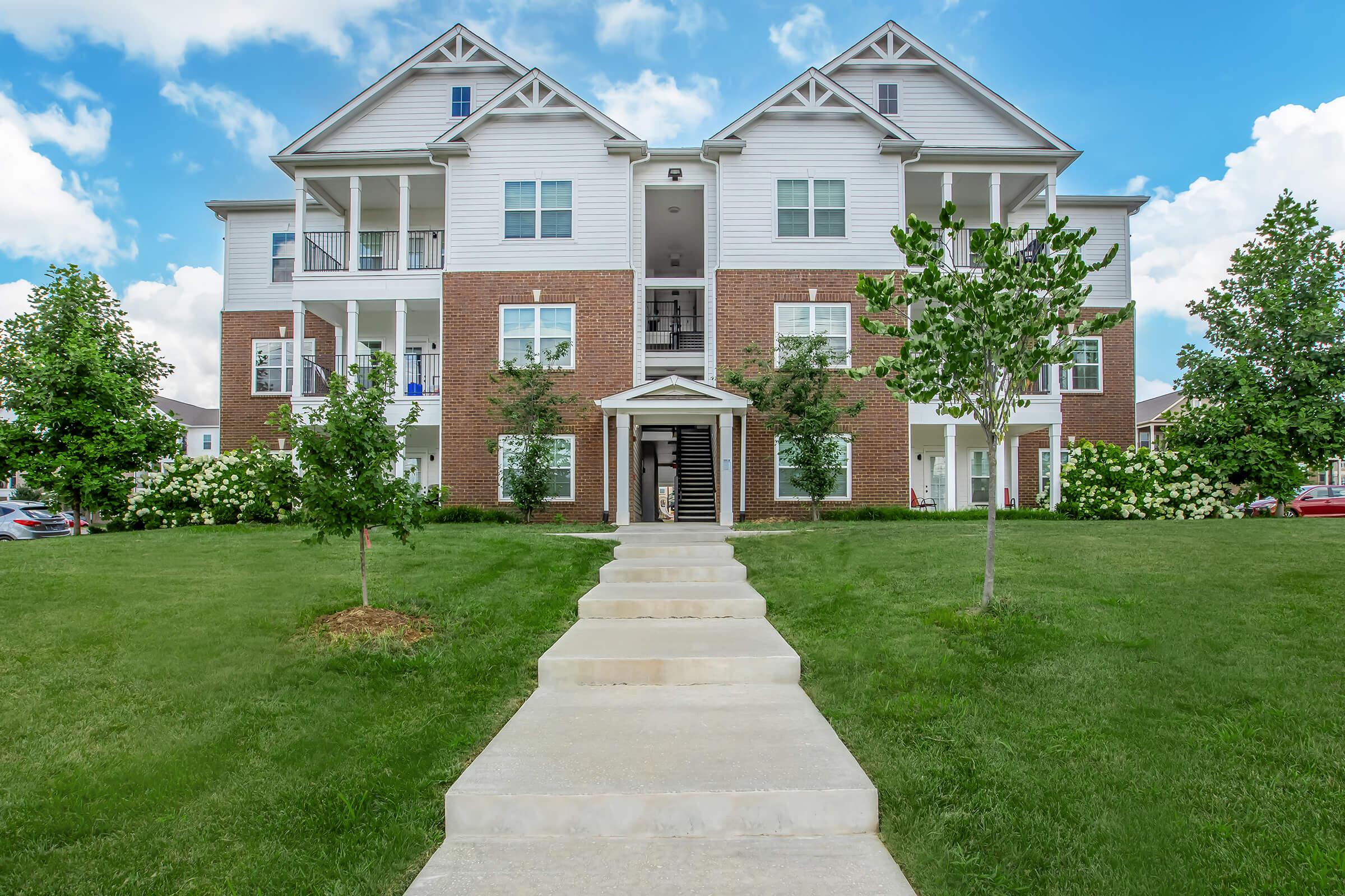 A well-maintained apartment building with a brick exterior, featuring multiple balconies and surrounded by lush green grass and small trees. A concrete walkway leads to the entrance, which has steps and a covered porch area. The sky is partly cloudy, adding to the bright atmosphere.