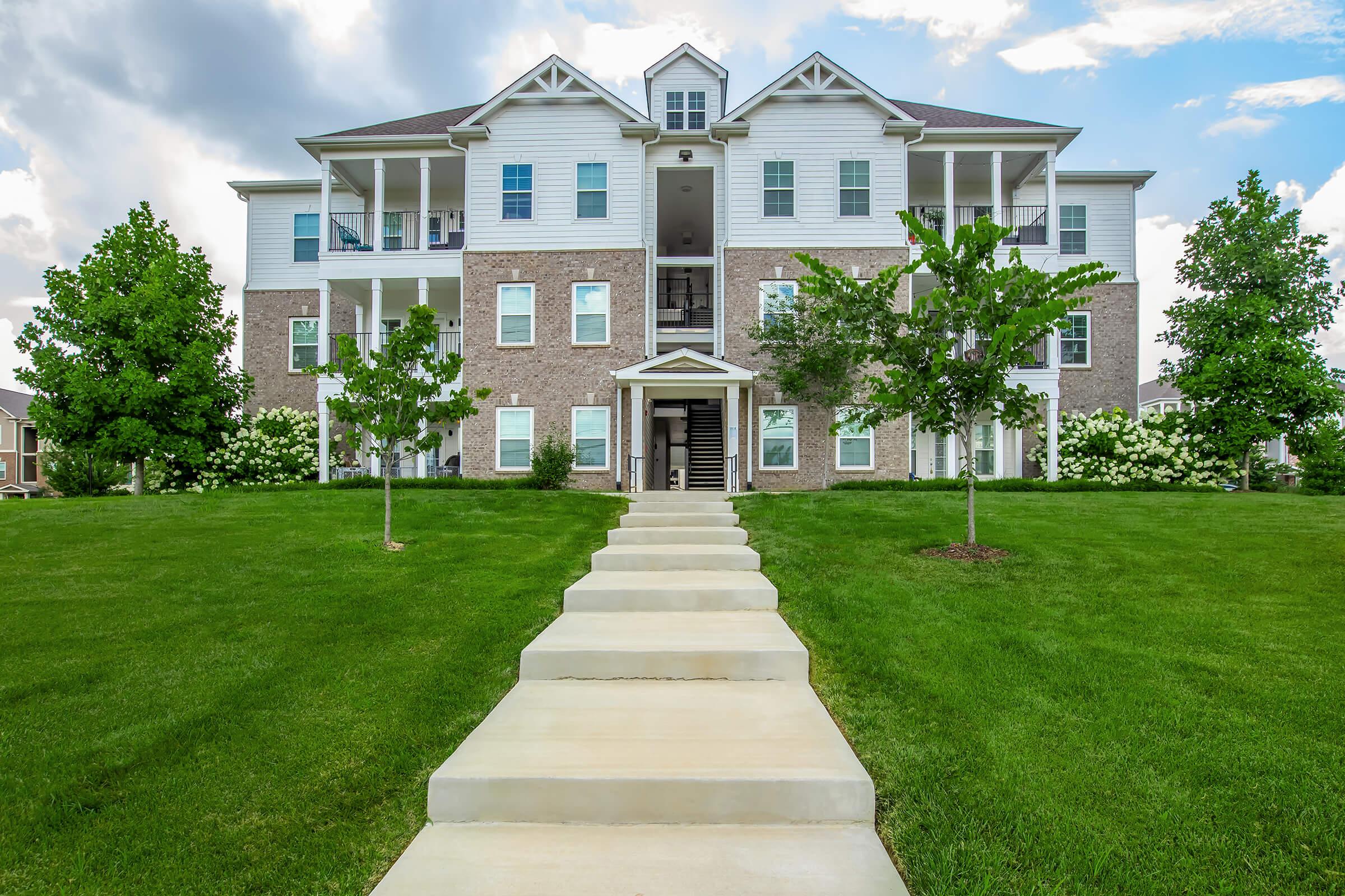 A modern three-story residential building with brick and siding, featuring balconies and stairways. The structure is surrounded by a well-maintained lawn with green grass and small trees, under a partly cloudy sky. A concrete pathway leads from the foreground to the entrance of the building.
