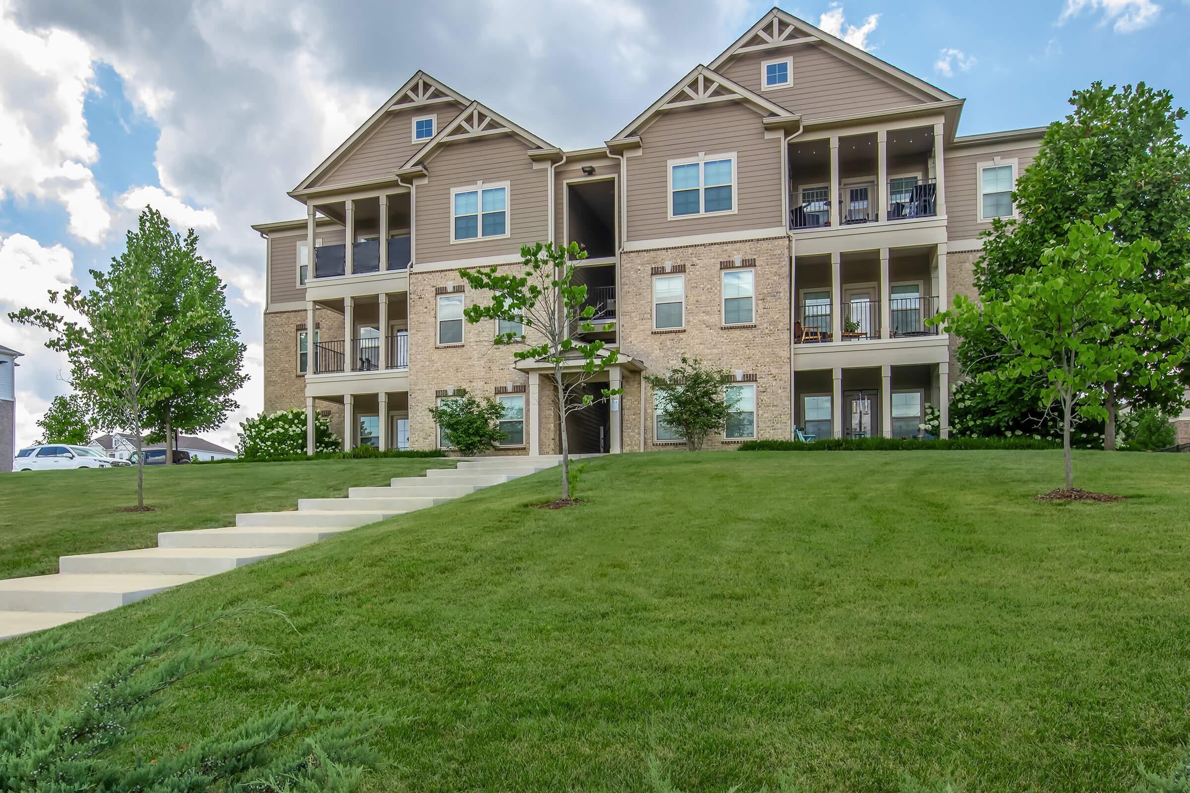 A multi-unit residential building with a tan facade, featuring balconies on each level. The building is surrounded by freshly mowed green grass and small trees, with a concrete pathway leading up to the entrance. The sky is partly cloudy, adding a serene atmosphere to the scene.