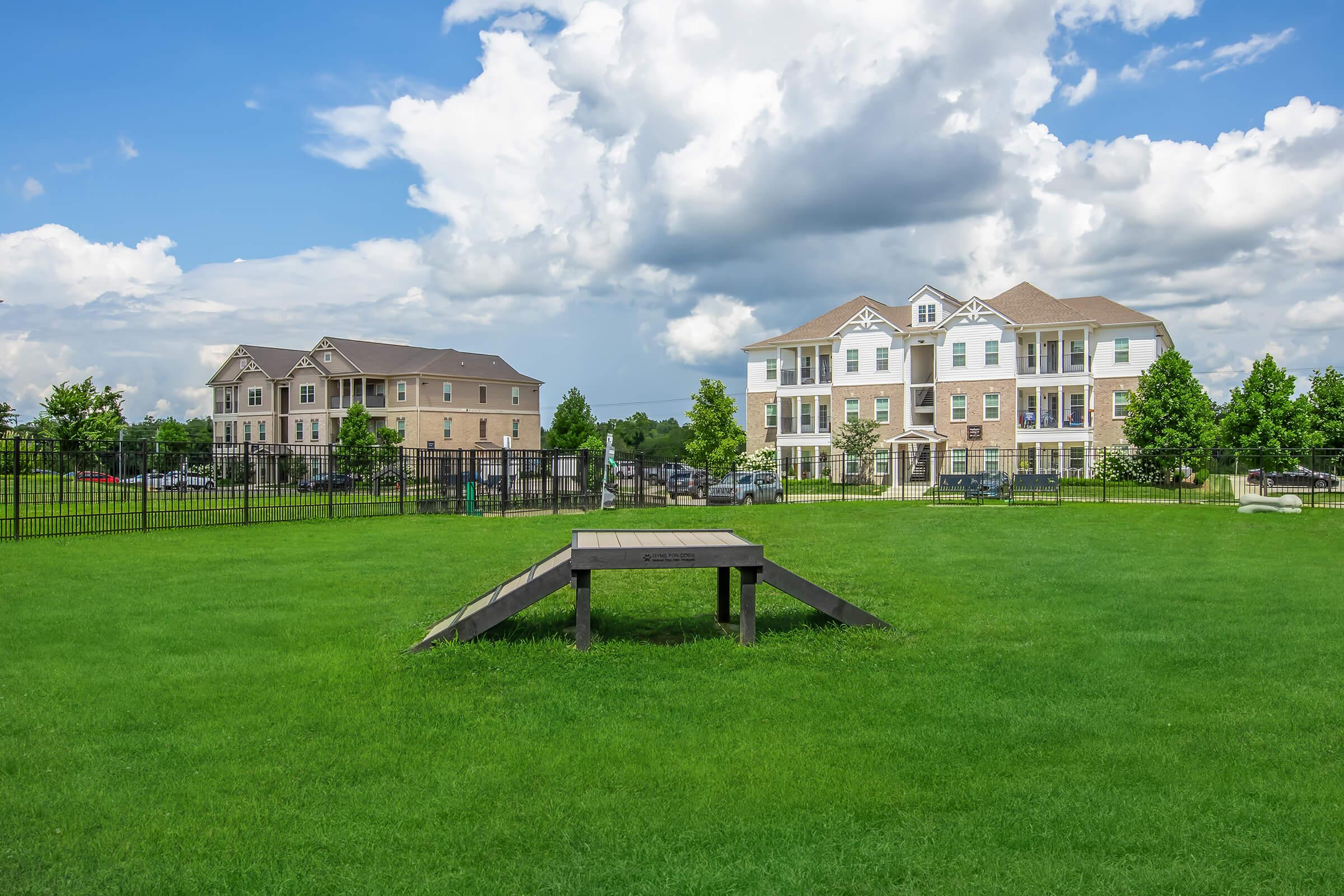A grassy area featuring a wooden platform and two multi-story residential buildings in the background. The sky is partly cloudy, with a mix of blue and white clouds. A fenced area is visible, suggesting a well-maintained community space.