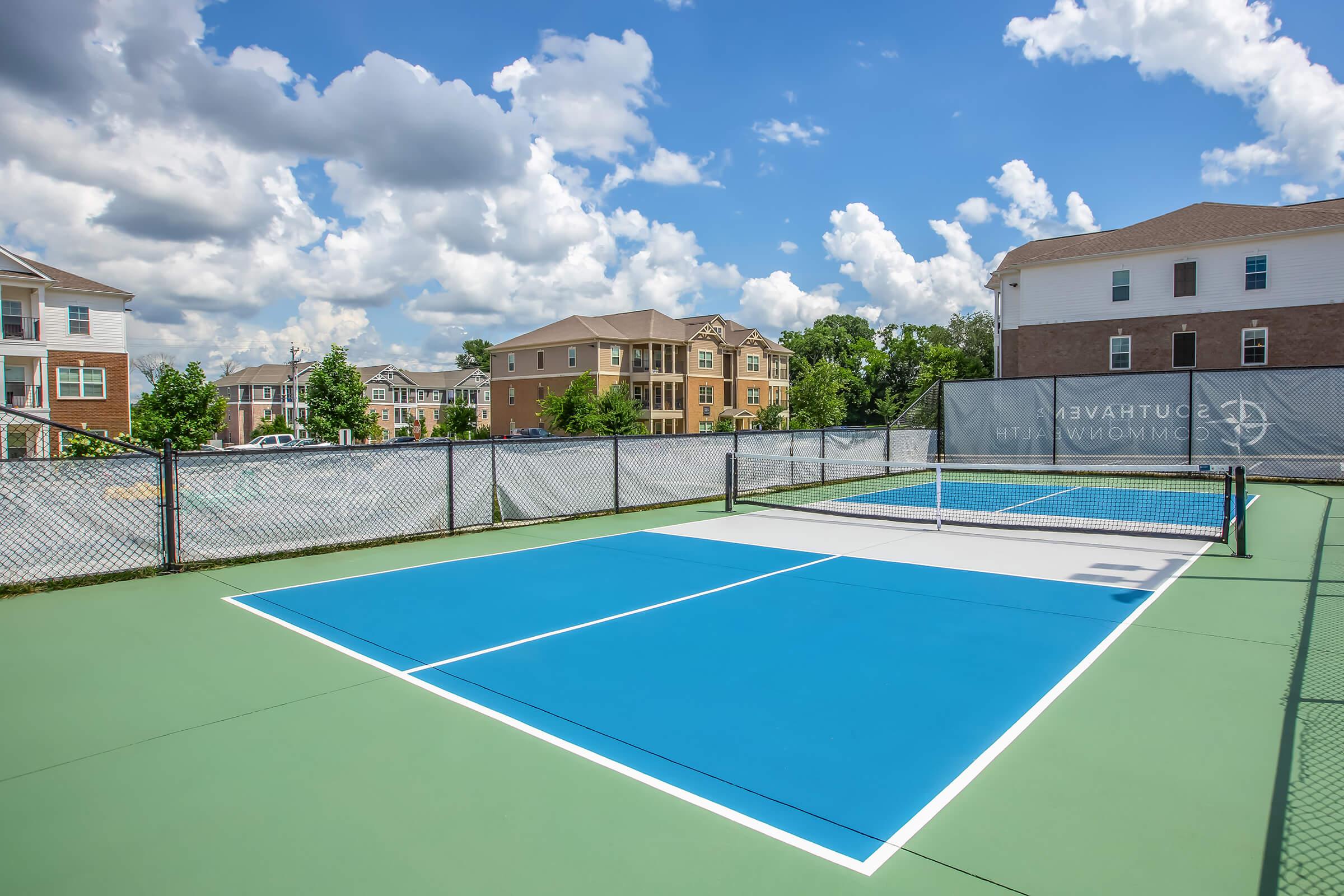 A brightly colored tennis court with blue and green surfaces, surrounded by a fence. In the background, there are residential buildings under a partly cloudy sky, creating a vibrant and inviting atmosphere for outdoor activities.