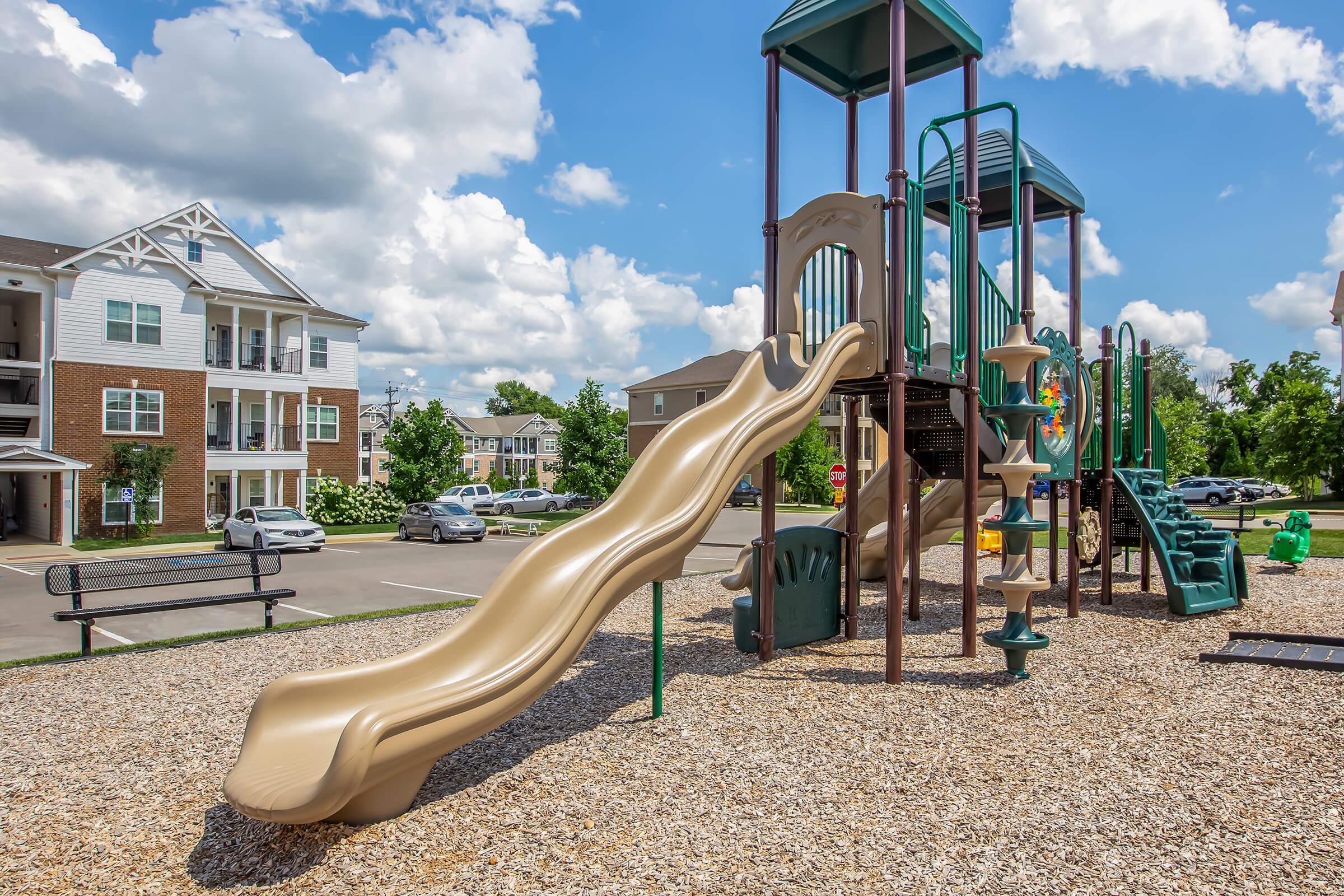 A playground featuring a large golden slide, climbing structures, and various play elements, surrounded by gravel. In the background, residential buildings and parked cars are visible under a bright blue sky with fluffy white clouds. Trees add greenery to the scene.