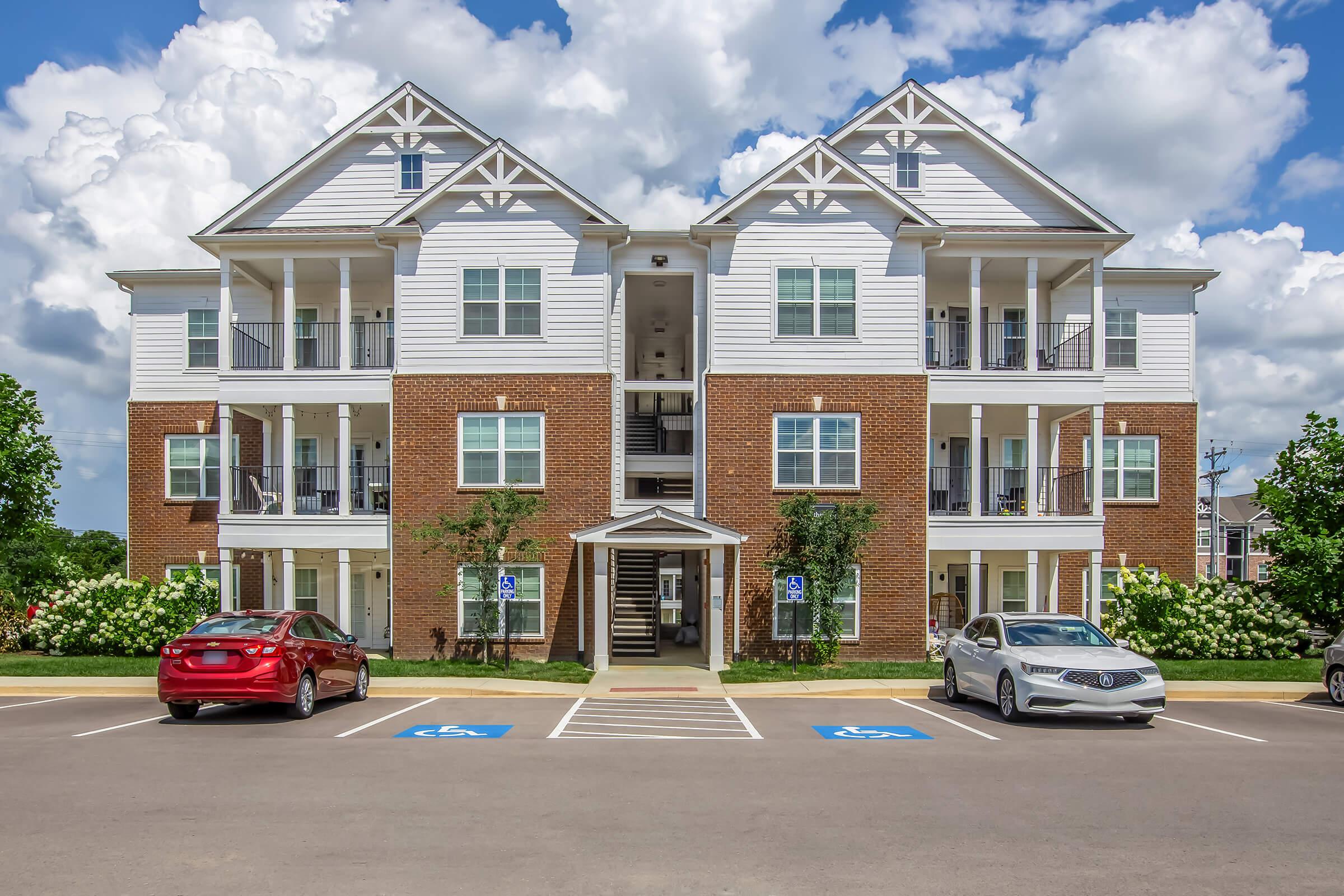 A modern three-story apartment building with a brick exterior and white siding. The building features multiple balconies and a staircase in the center. There are two parked cars in the handicapped parking spaces in front, surrounded by green shrubs under a partly cloudy sky.