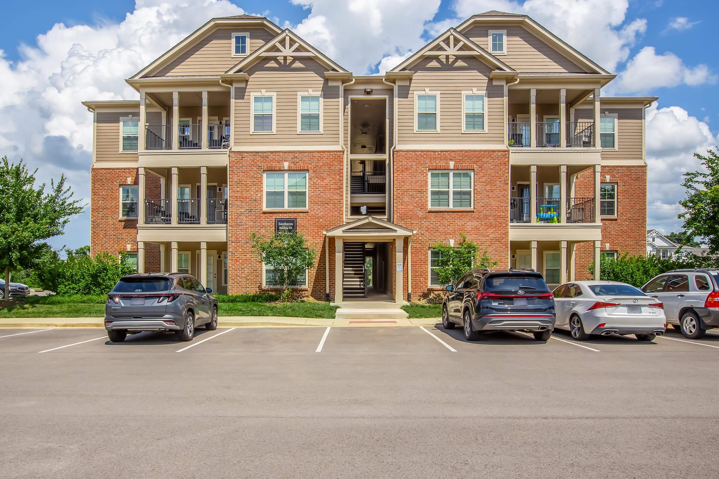 A three-story apartment building with a brick and beige exterior. It features balconies on each floor, with several parked cars in front and green landscaping on either side. The sky is partly cloudy, enhancing the bright atmosphere of the scene.