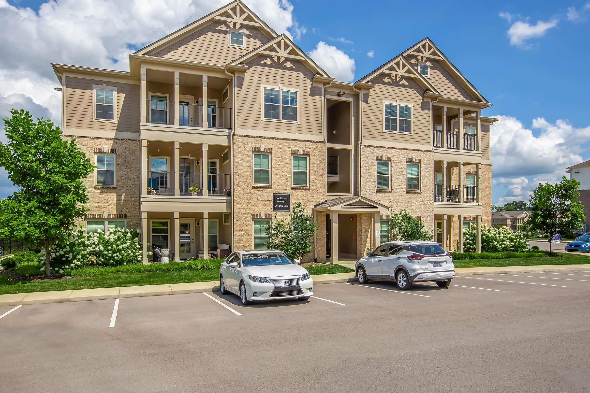 A modern, multi-story apartment building with balconies, surrounded by greenery and flowering shrubs. Two cars are parked in the lot in front of the building. The sky is blue with fluffy clouds, creating a bright and inviting atmosphere.