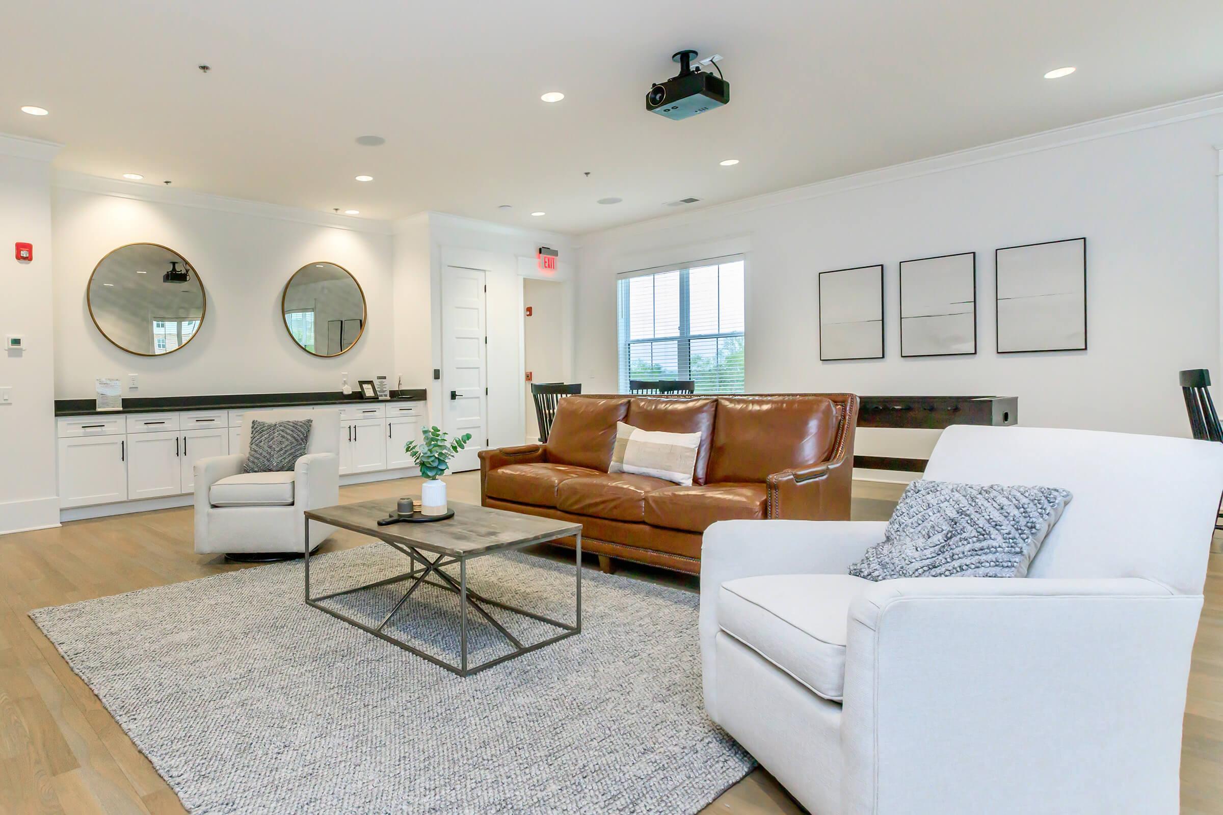 A modern living area featuring a brown leather sofa, two white armchairs, and a coffee table on a large area rug. The room has two round mirrors on the wall, a projector mounted on the ceiling, and large windows letting in natural light, creating a bright, inviting atmosphere.